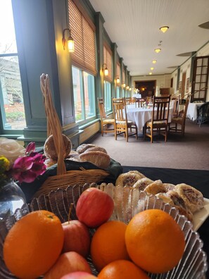 A dining area with tables and chairs, a buffet table with fruits and pastries, and large windows with blinds.