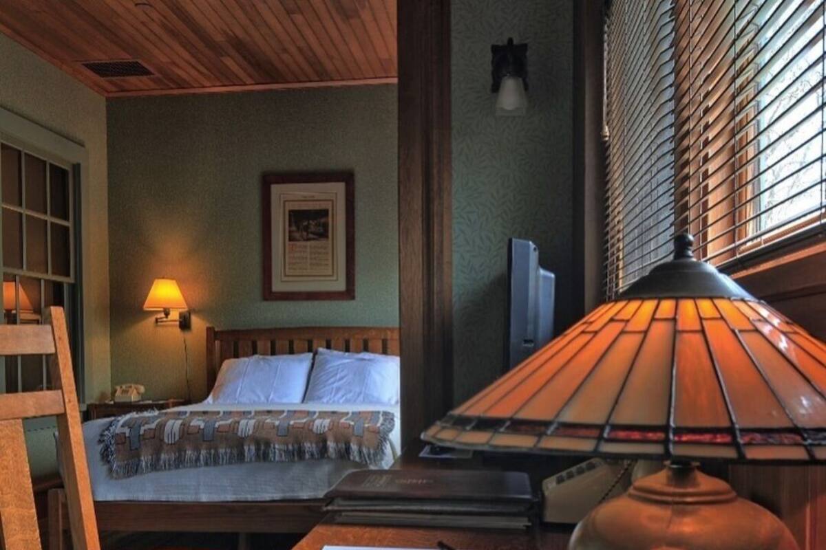 A hotel room with a wooden bed, a desk with a lamp, a chair, and a framed picture on the wall.