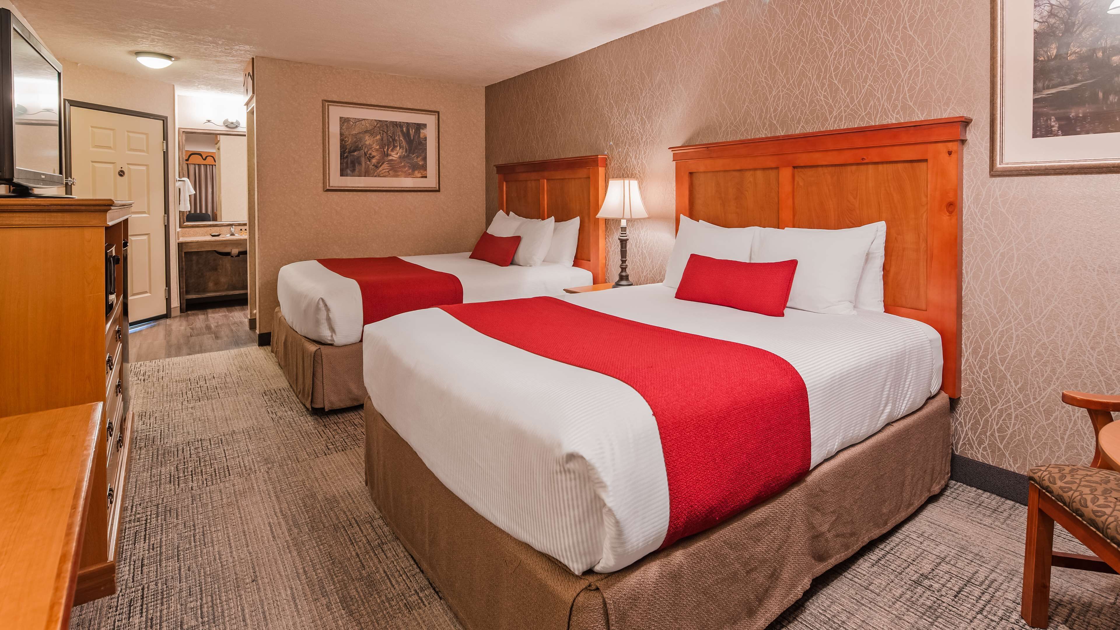 A hotel room with two beds, a wooden headboard, a red and white bedspread, a wooden dresser, a mirror, and a framed picture on the wall.