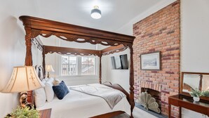 A four-poster bed with a brick fireplace, a bedside table with a lamp, and a window with a view of buildings. - Grenoble House (New Orleans)