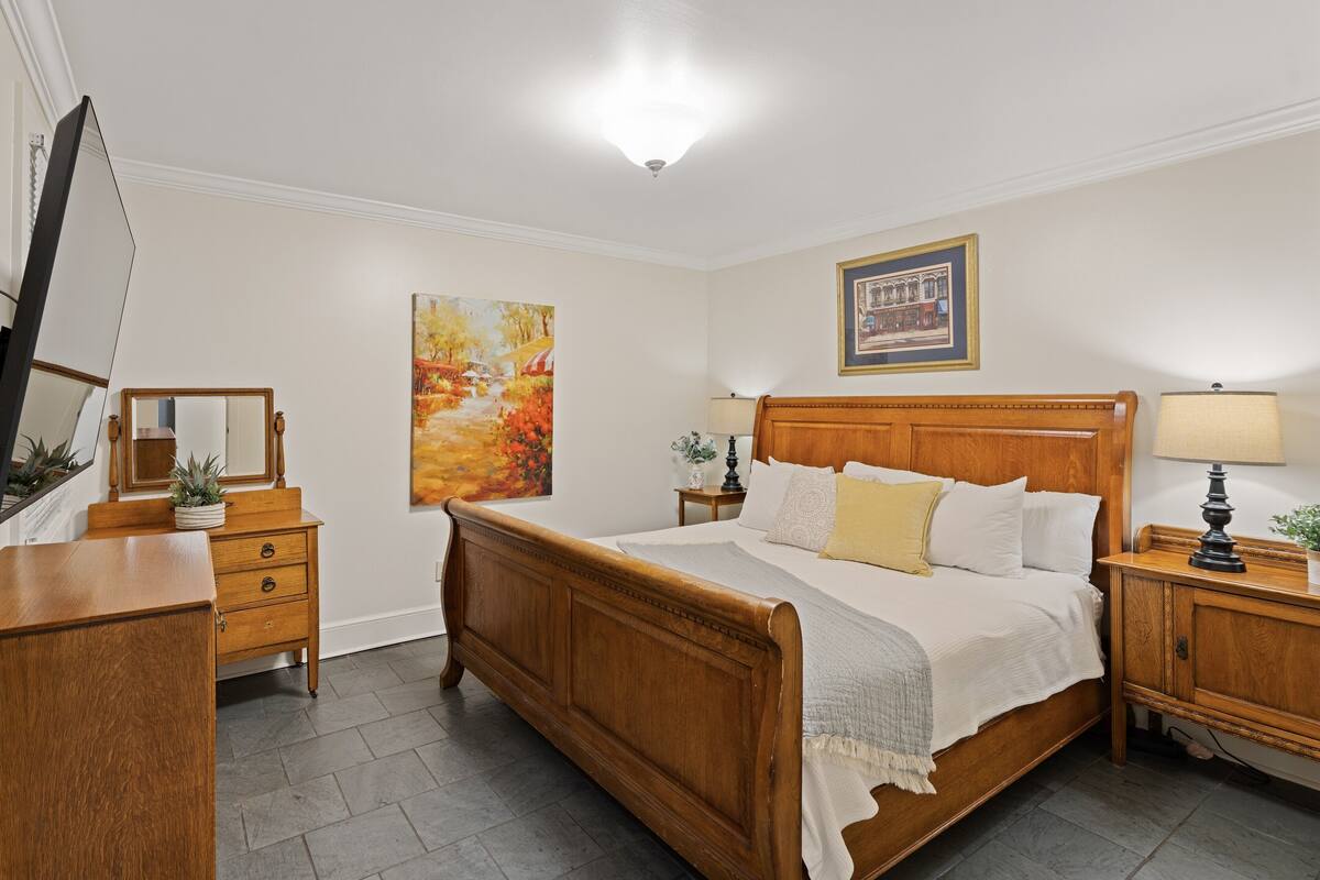 A bedroom with a wooden bed, a nightstand with a lamp, a dresser, a mirror, and a painting on the wall.