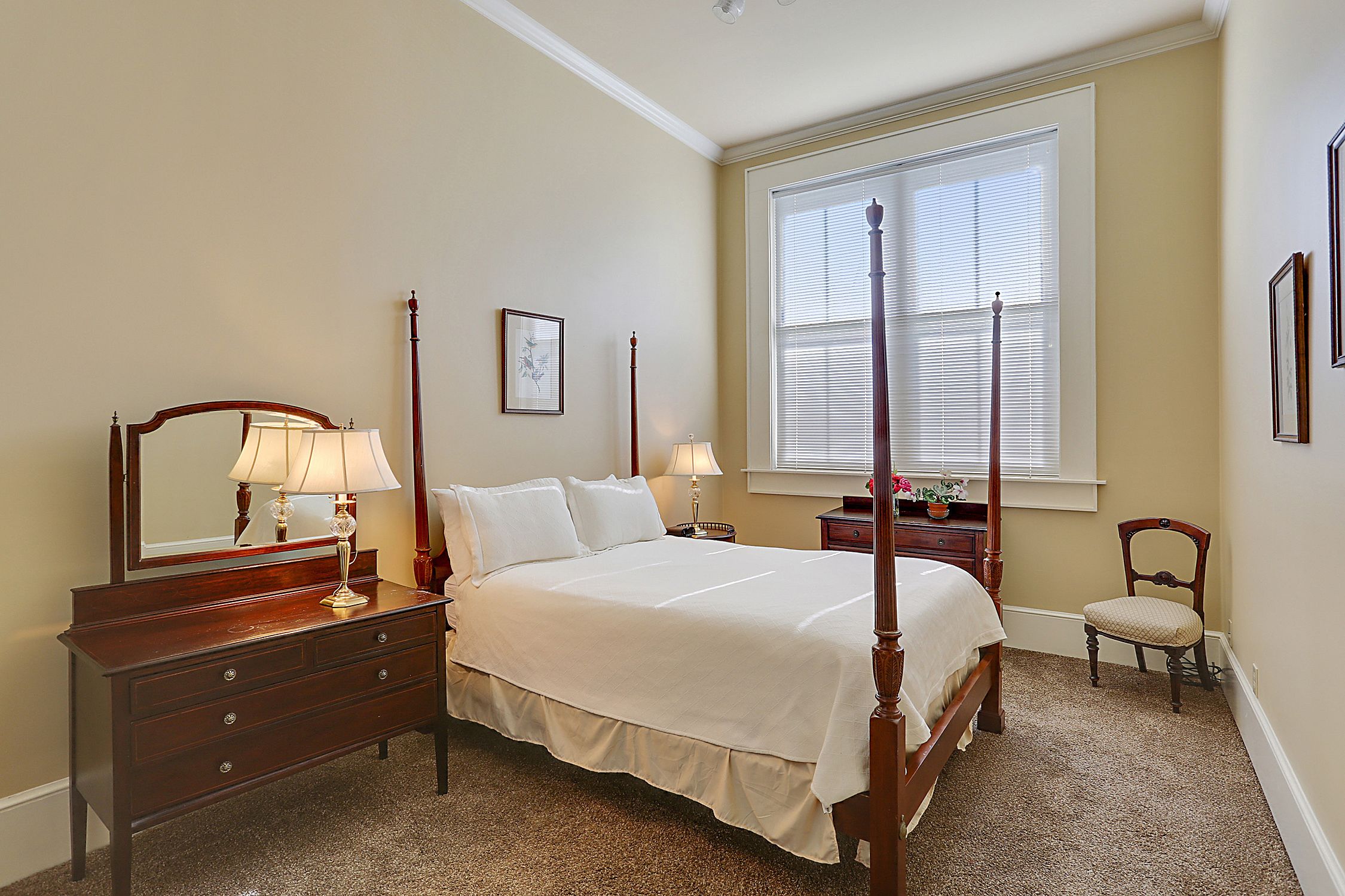A bedroom with a four-poster bed, a wooden dresser with a mirror, two bedside tables with lamps, and a chair.