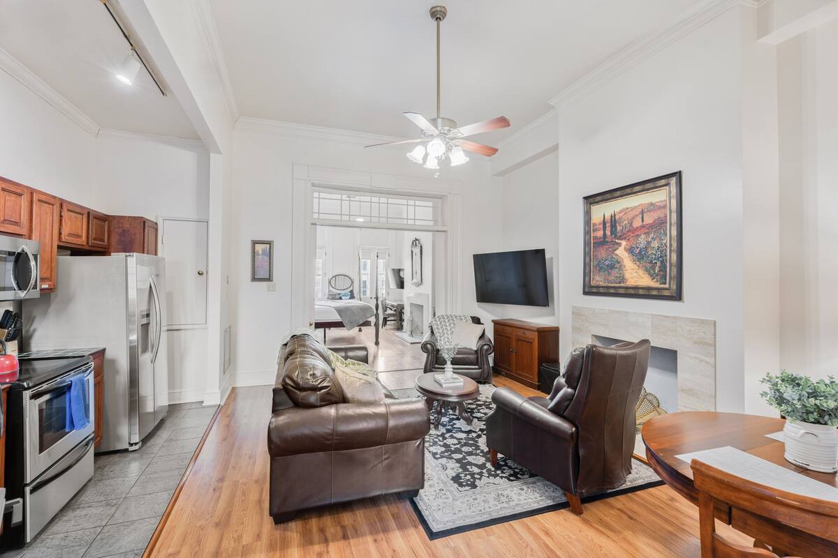 A modern living room with a leather sofa, a coffee table, and a fireplace. There is a kitchen area with appliances and wooden cabinets.