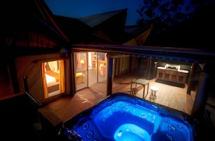 A hot tub area with a wooden deck, a glass door leading to a room with a lit lamp, and a bar area with stools.