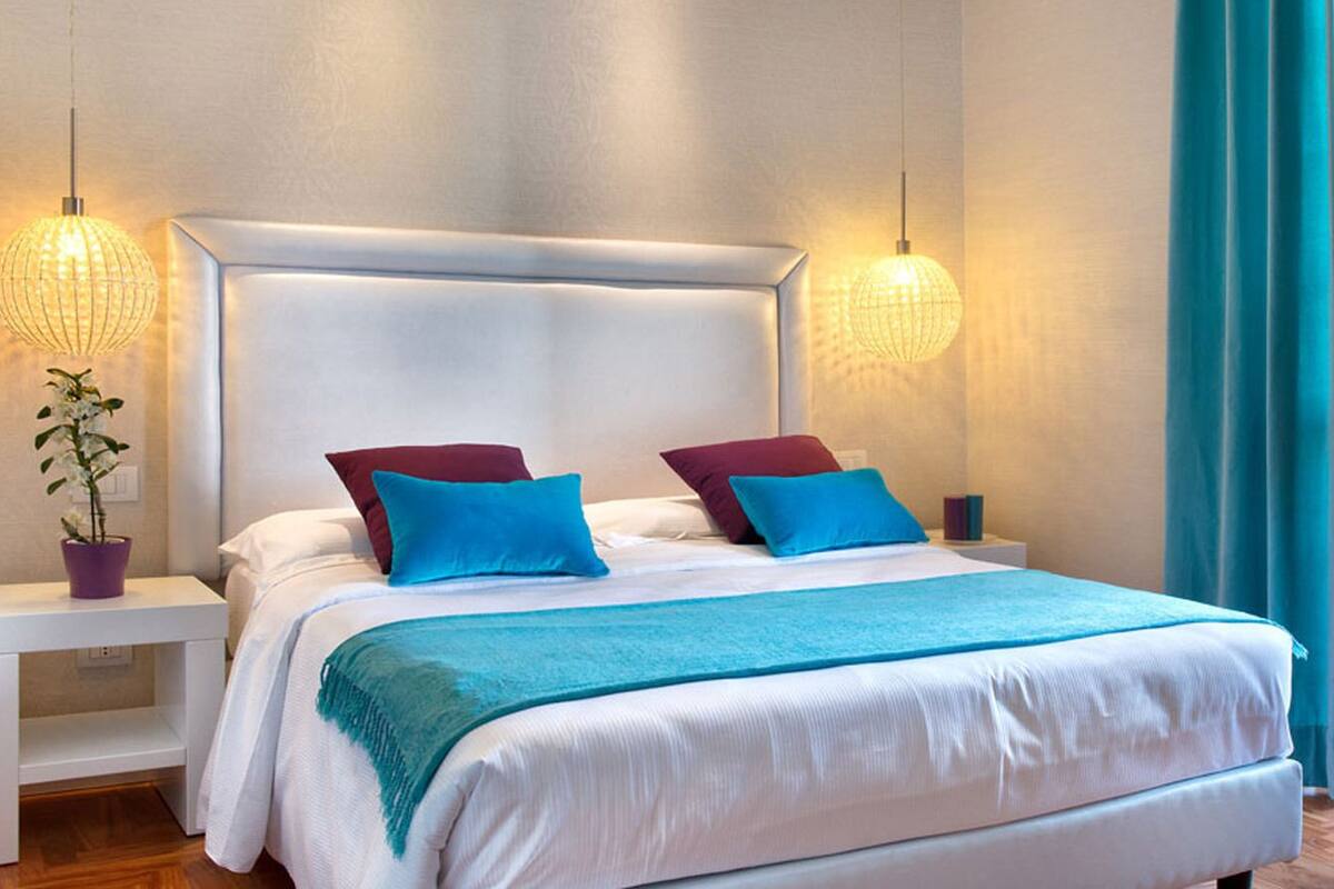 A modern bedroom with a large bed, two nightstands, a potted plant, and a view through a window with curtains.