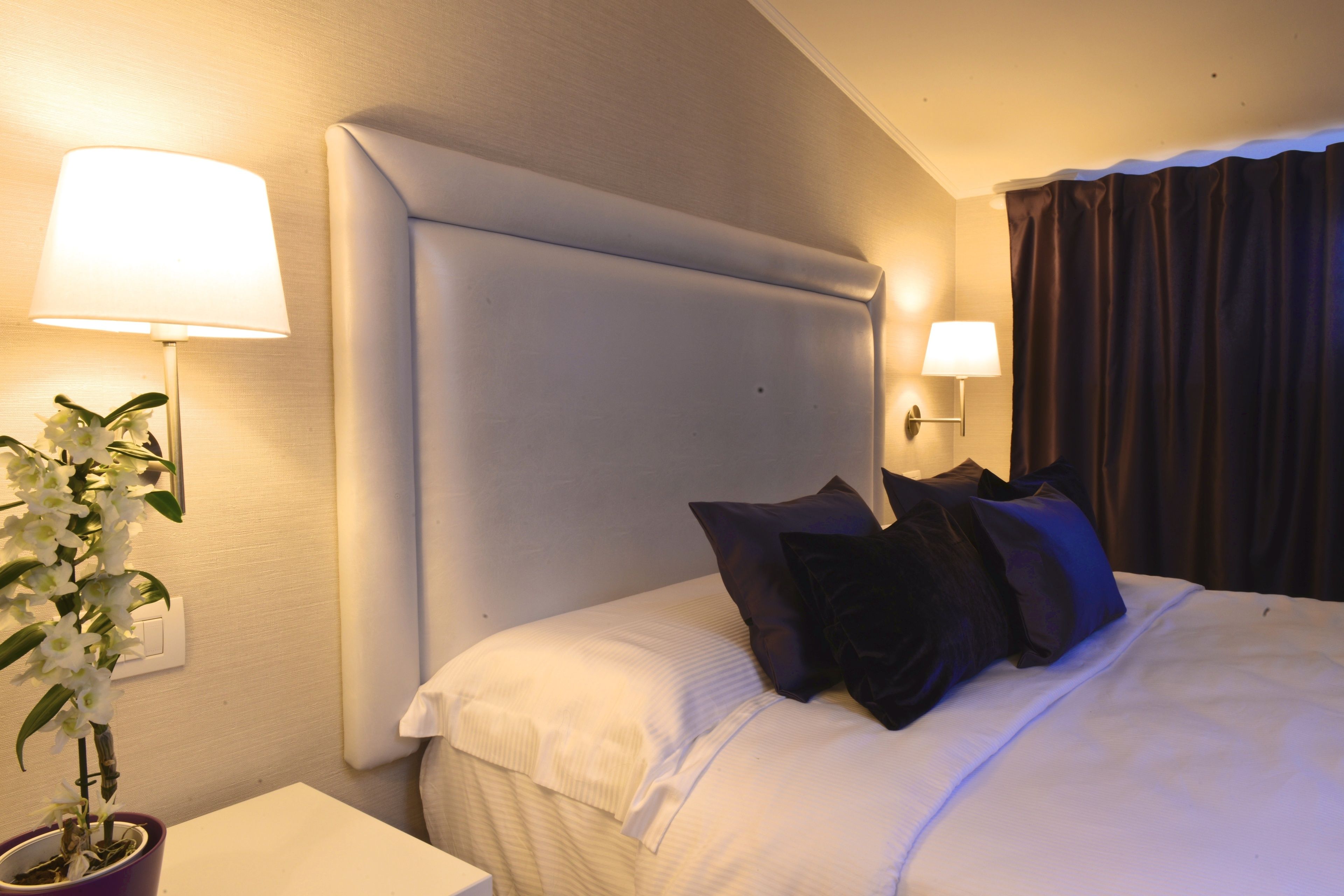 A hotel room with a large bed, two bedside lamps, a potted plant, and a headboard.