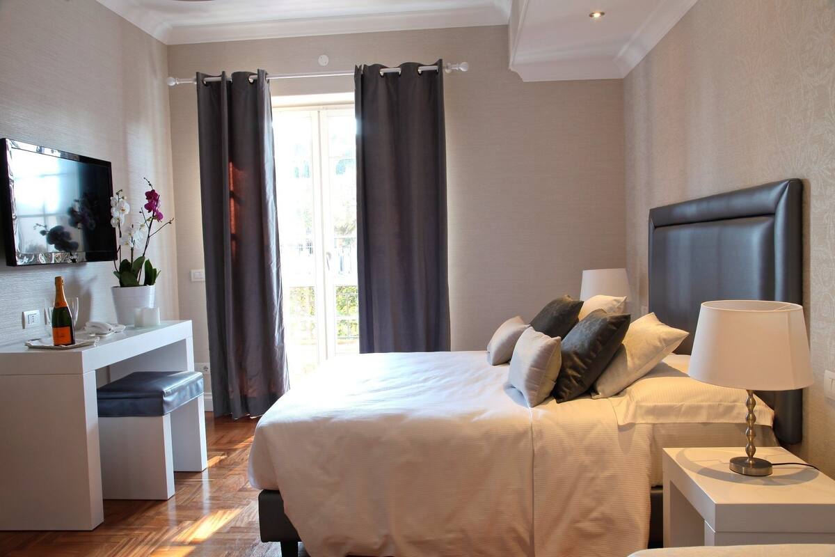A hotel room with a bed, bedside table, TV, and a small desk with a bottle and flowers.