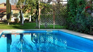 A rectangular swimming pool with a clear blue water surface, surrounded by a wooden deck and a lush green garden with various plants and trees. - La Villetta Suite (Ciampino)