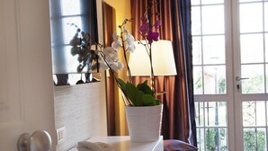 A hotel room with a potted orchid plant on a bedside table, a lamp, and a view of the outside through a window with curtains. - La Villetta Suite (Ciampino)
