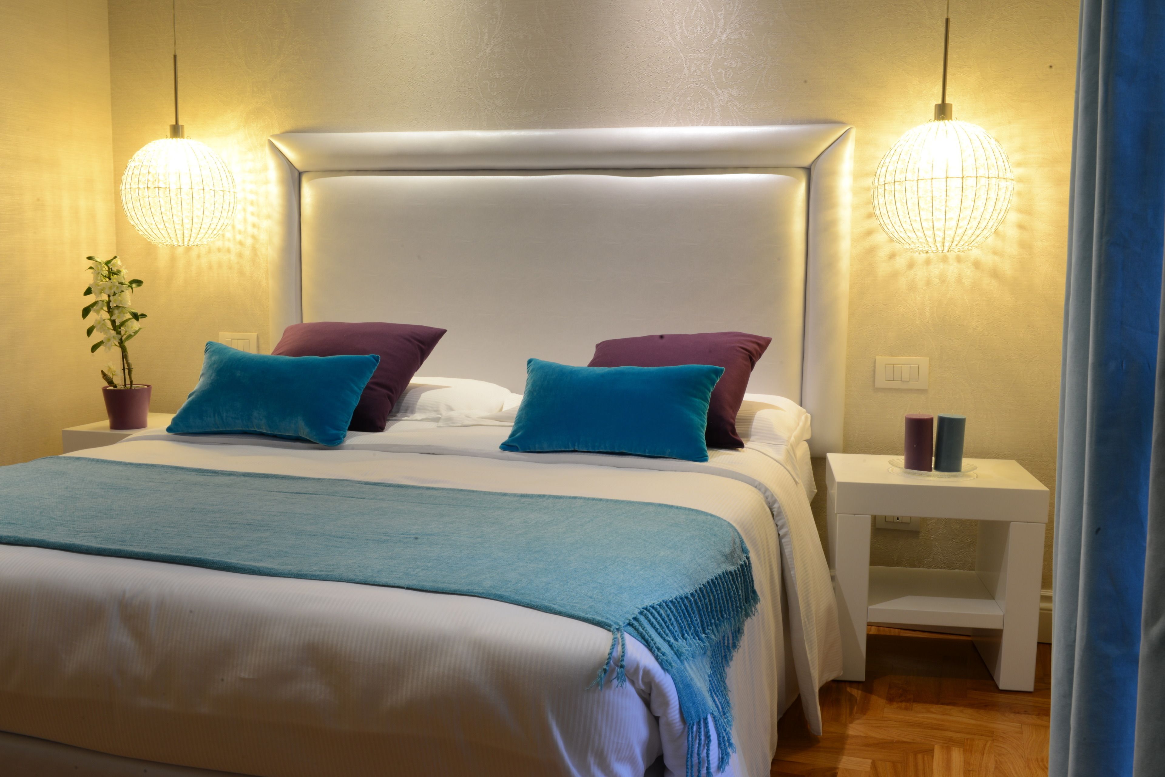 A neatly made bed with a white headboard, two bedside tables, and a potted plant on the left.