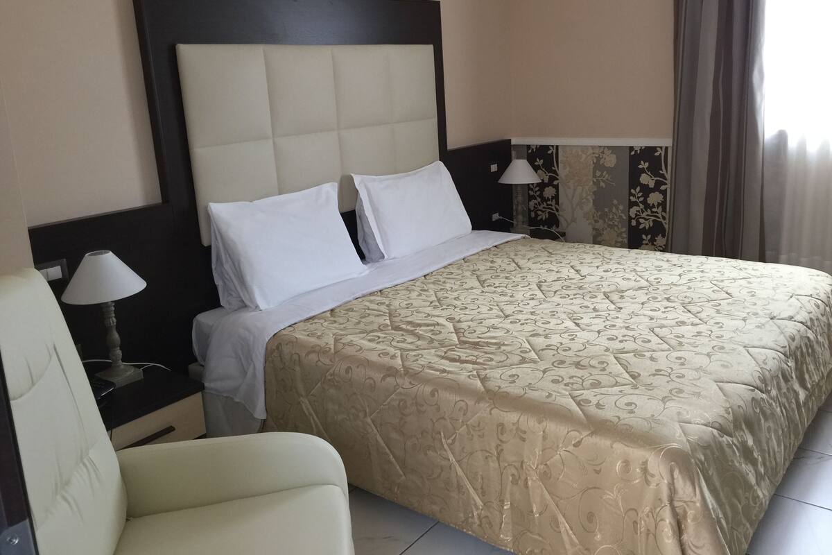 Ein Hotelzimmer mit einem großen Bett, einem Nachttisch mit Lampe, einem Sessel und einem Fenster mit Vorhängen.