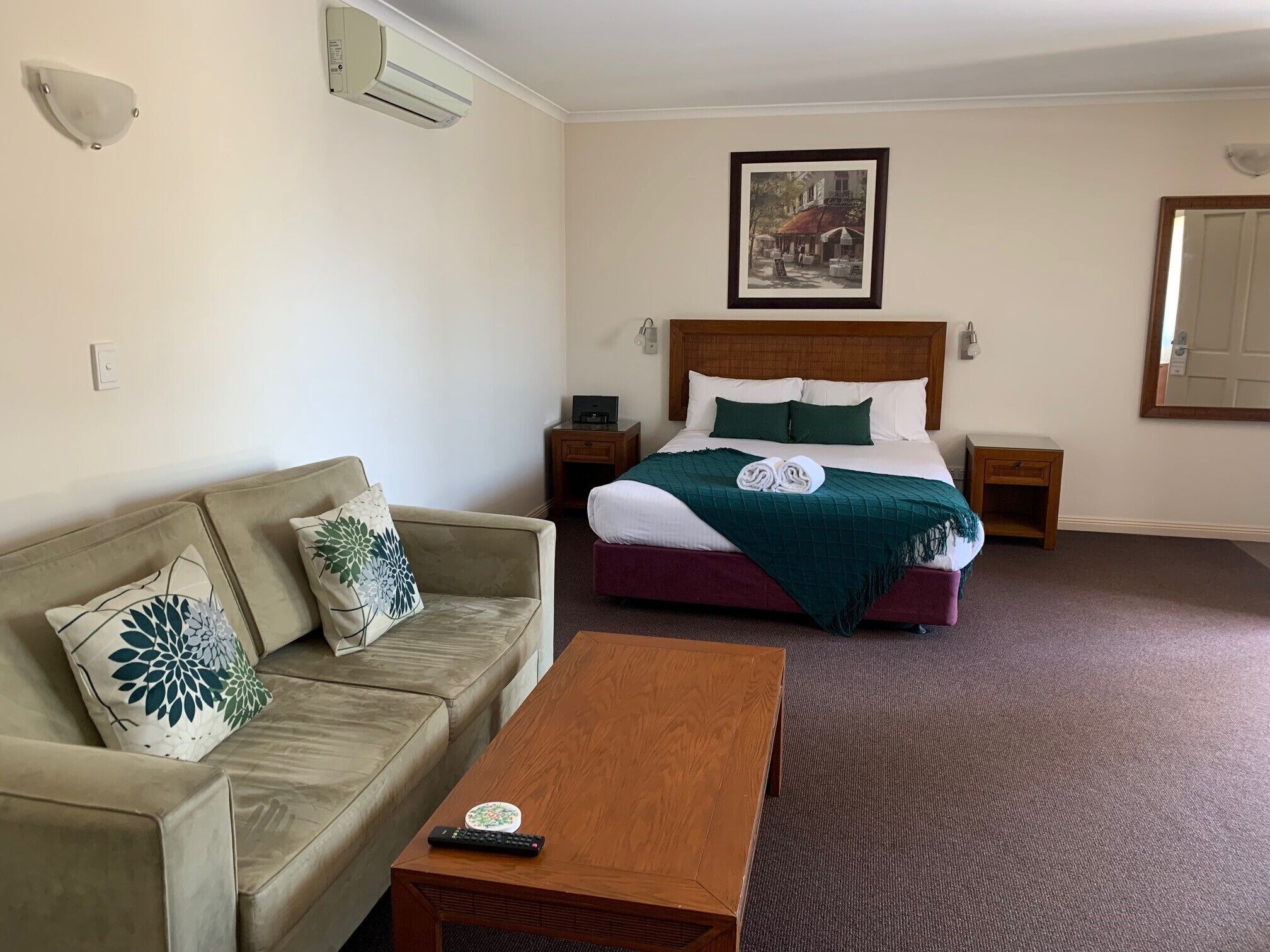 A hotel room with a bed, a sofa, a coffee table, a picture on the wall, and an air conditioning unit.