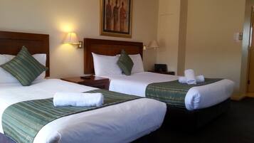 A hotel room with two single beds, each with a green-striped bedspread, white pillows, and a wooden headboard. There are bedside tables with lamps, and a painting above the beds.