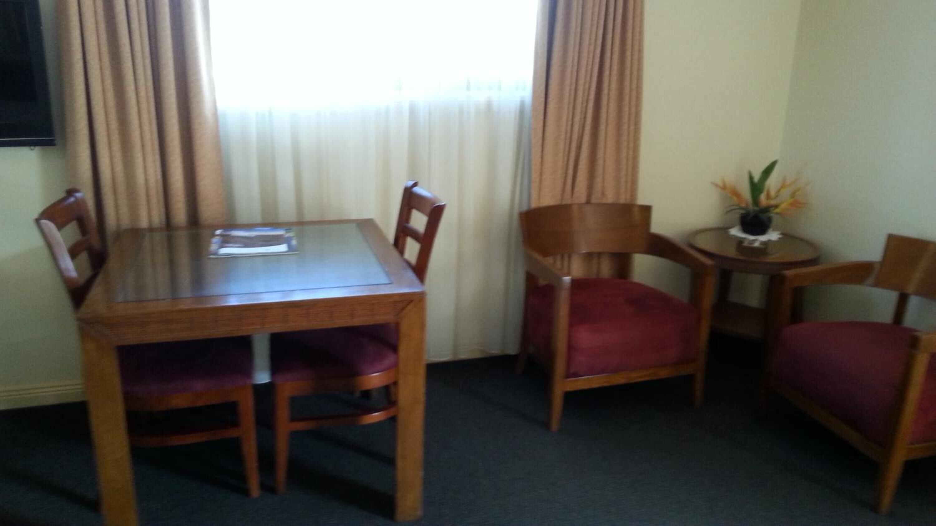 A hotel room with a wooden dining table, four chairs, a small side table with a vase, and a window with curtains.