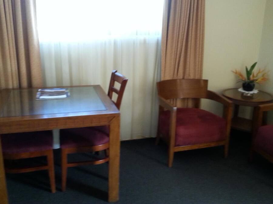 A hotel room with a wooden dining table, four chairs, a small side table with a vase, and a window with curtains.