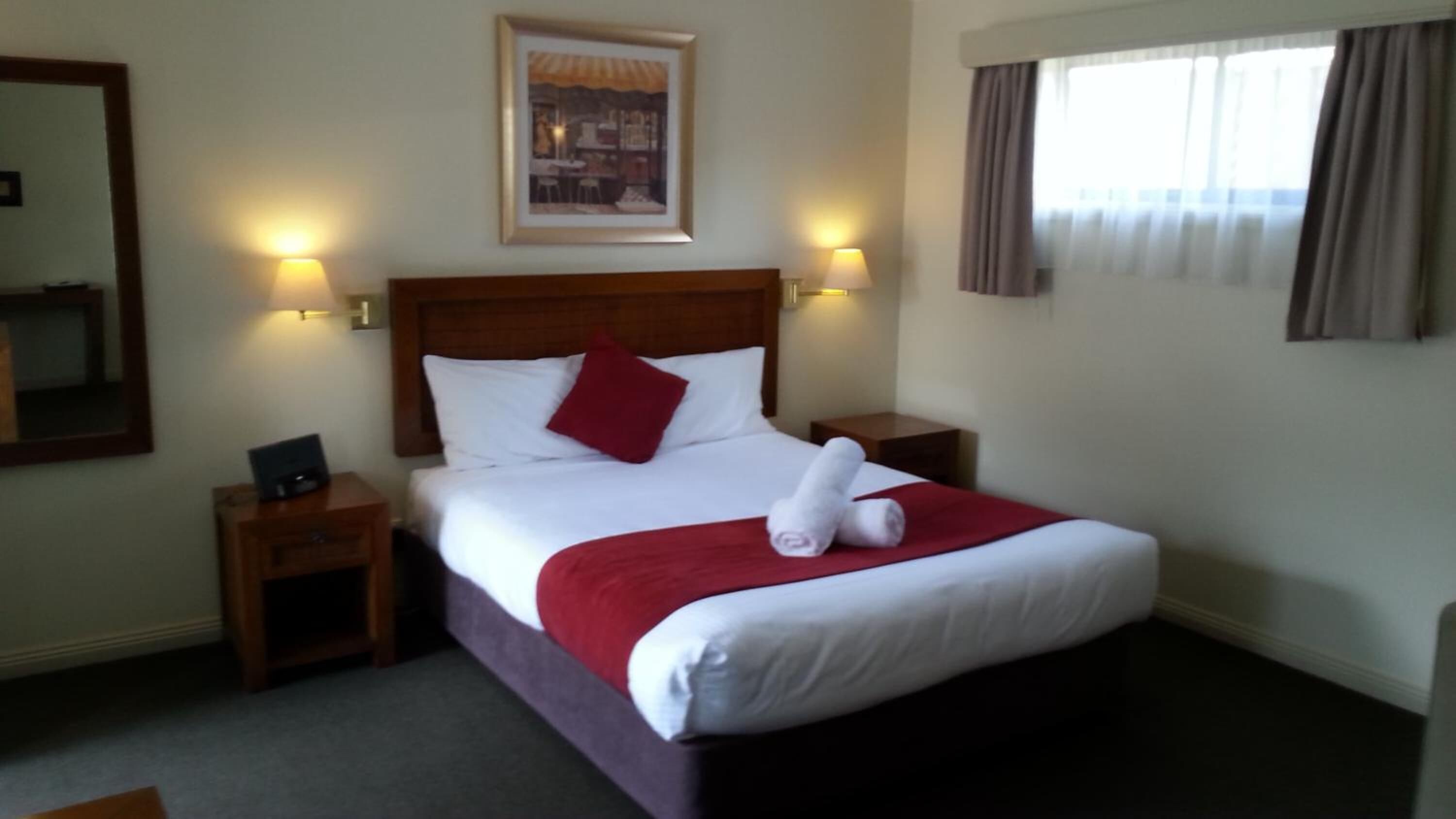 Une chambre d’hôtel avec un lit bien fait, deux tables de chevet, une commode, un tableau encadré au mur et une fenêtre avec des rideaux.