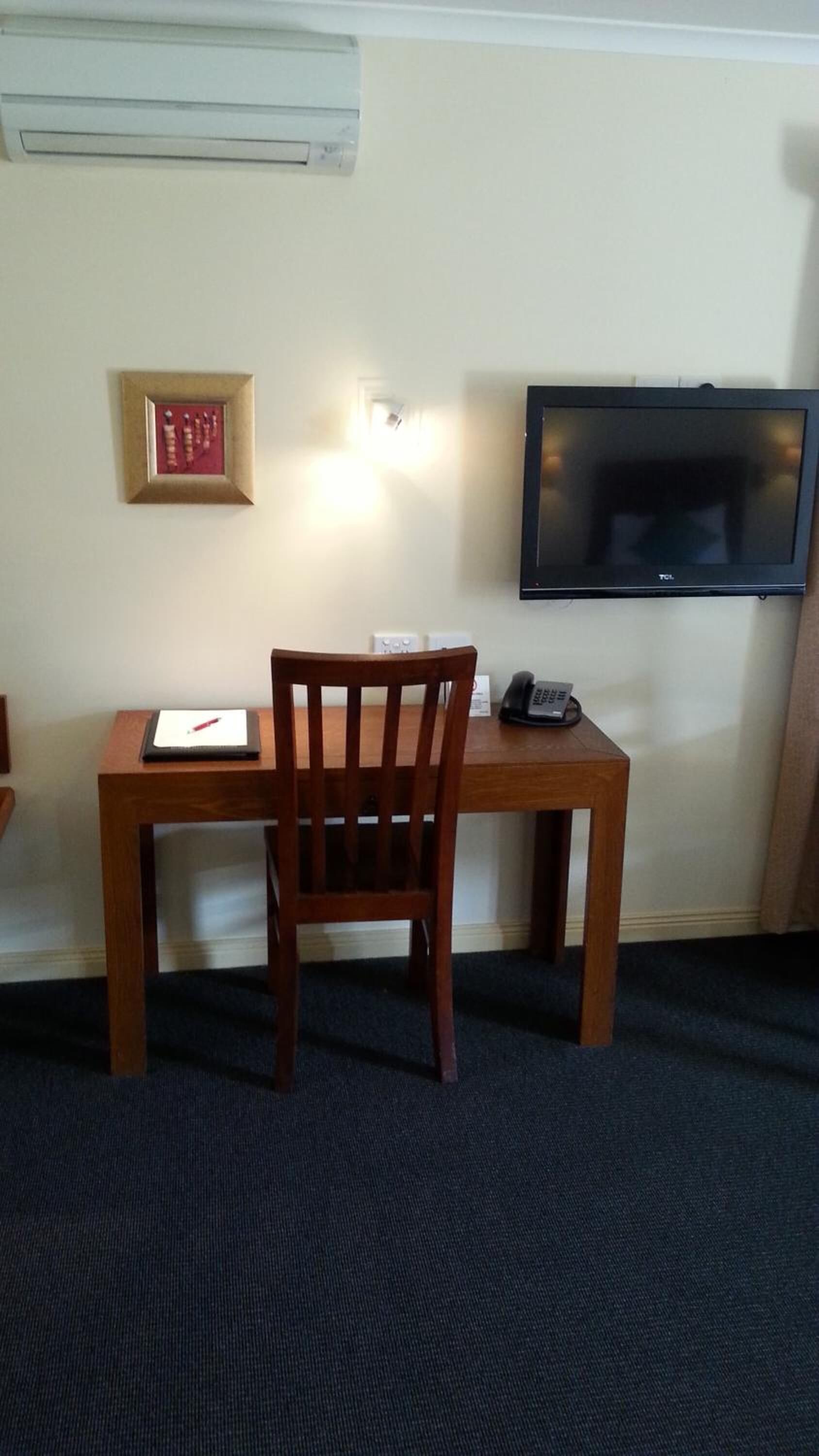 A room with a wooden desk, a chair, a framed picture, a telephone, and a television mounted on the wall.
