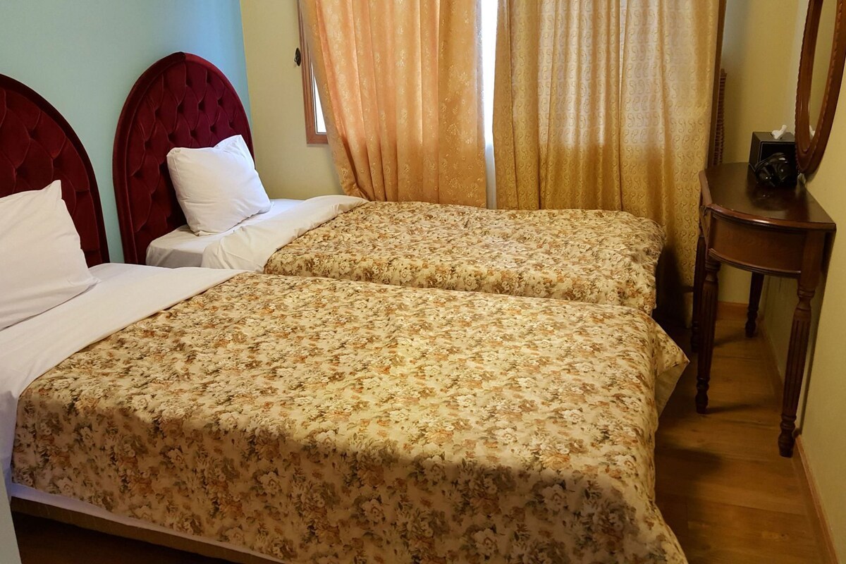 A double bed with floral bedding, a red upholstered headboard, and a wooden nightstand with a mirror.