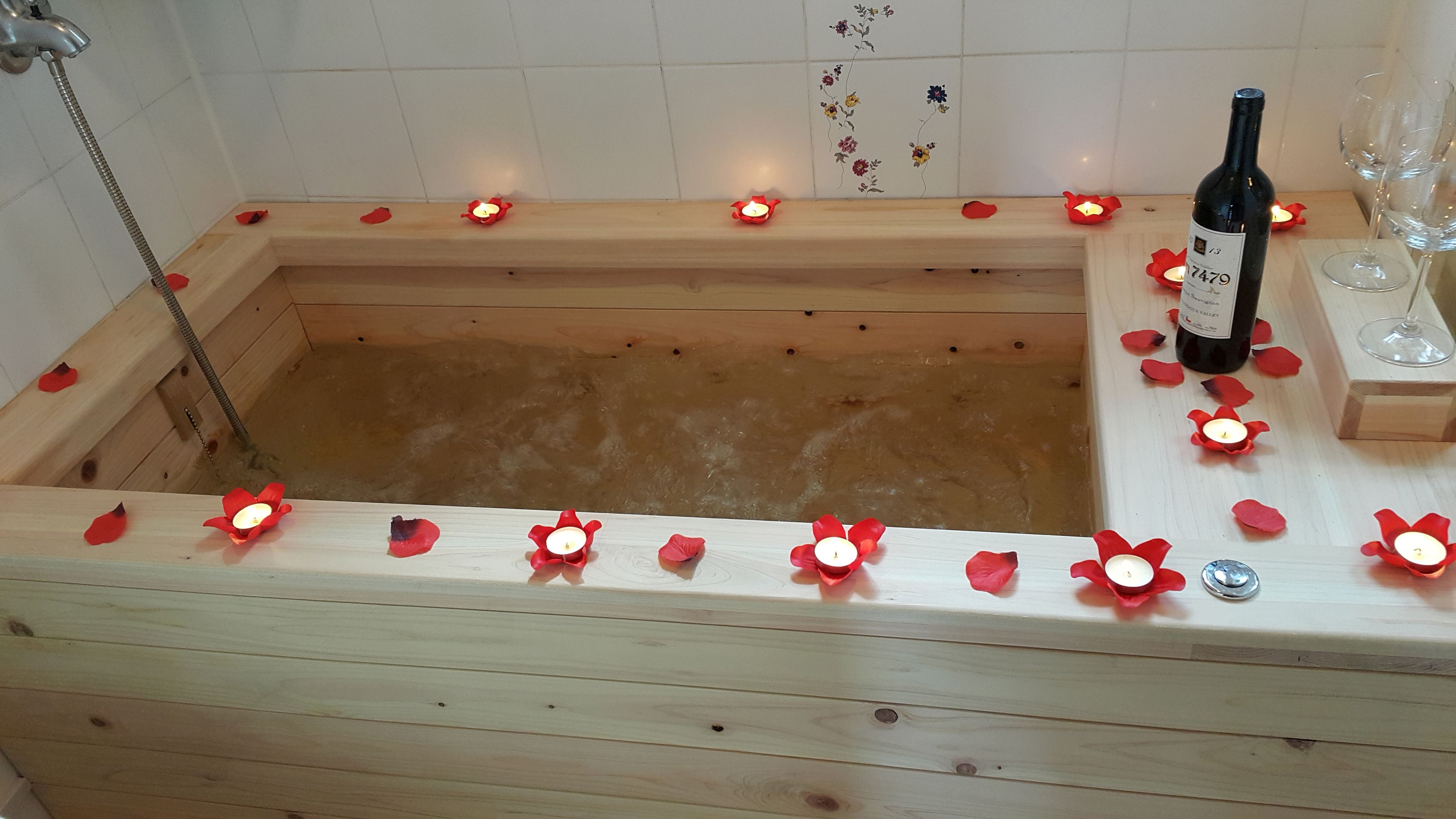A wooden bathtub with candles and rose petals, a bottle of wine, and glasses on a wooden table.