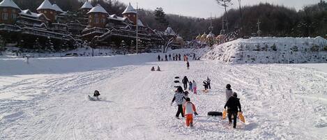 A snowy ski slope with people skiing and snowboarding. There are buildings with snow-covered roofs in the background.