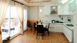 A kitchen with wooden ceiling beams, a dining area with a table and chairs, and a view of a balcony through a window with curtains.