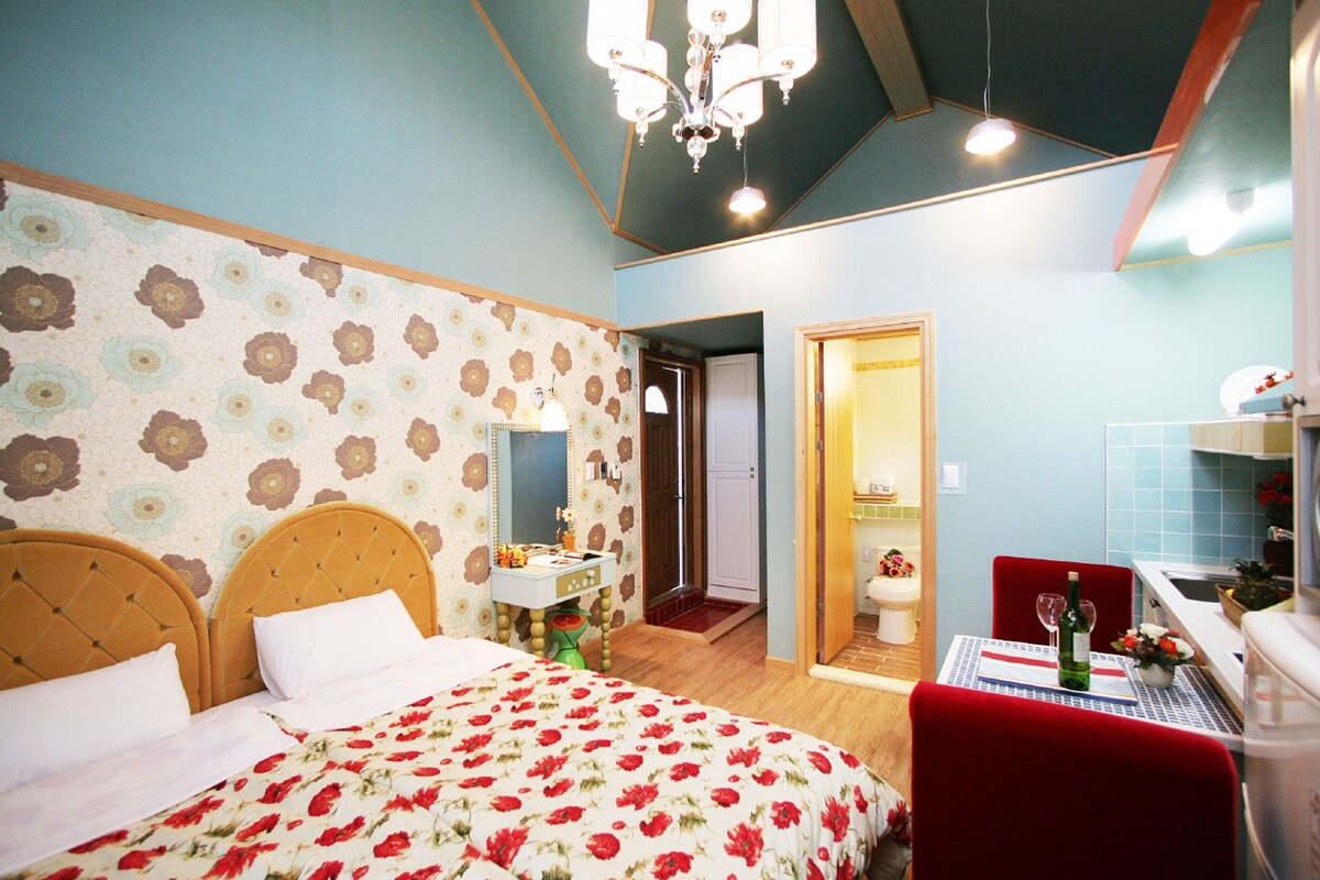 A bedroom with a floral-patterned bedspread, a wooden headboard, a nightstand, and a red chair.