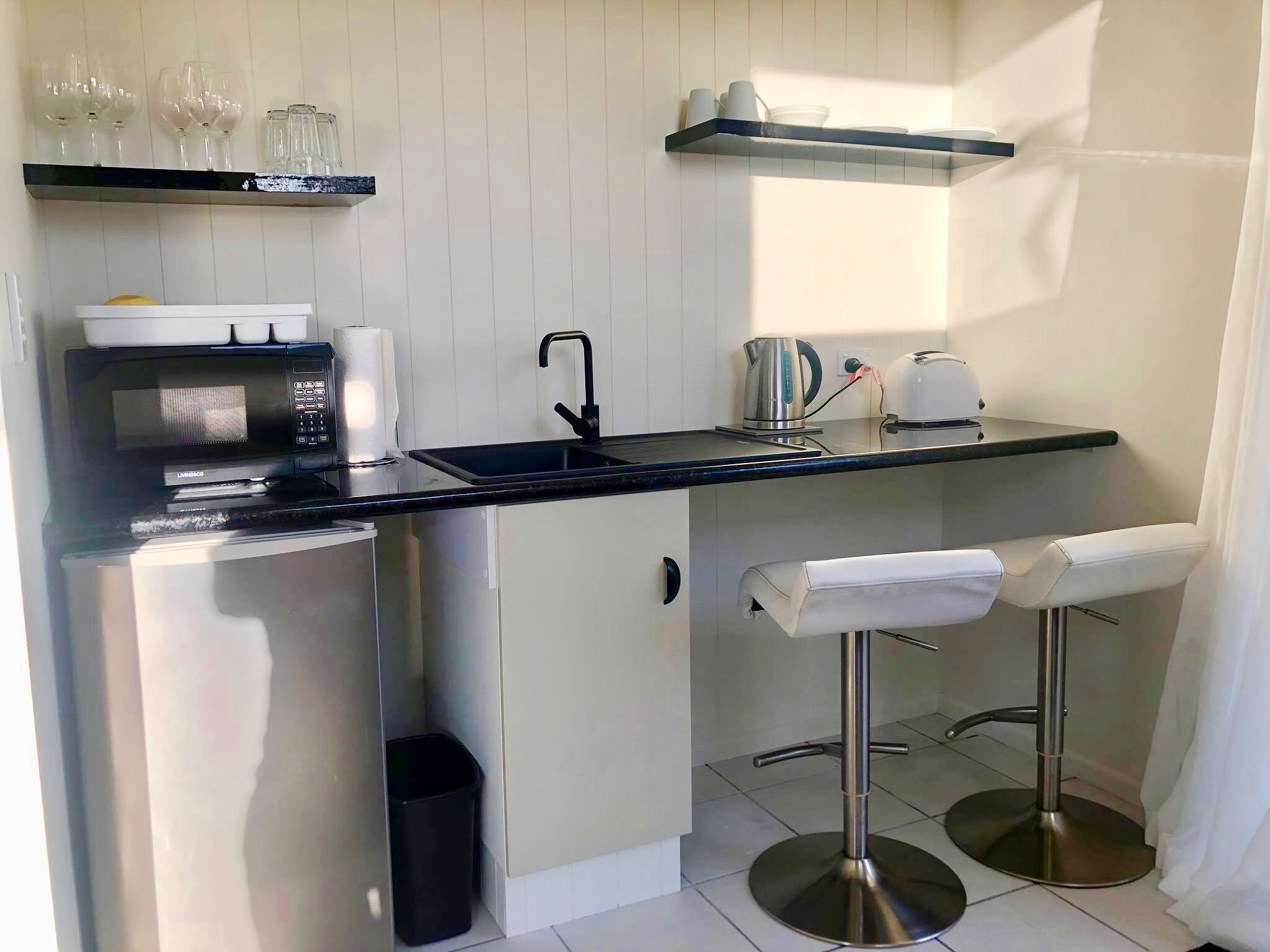 A compact kitchen with a microwave, toaster, kettle, and a sink. There are two bar stools and a trash bin.