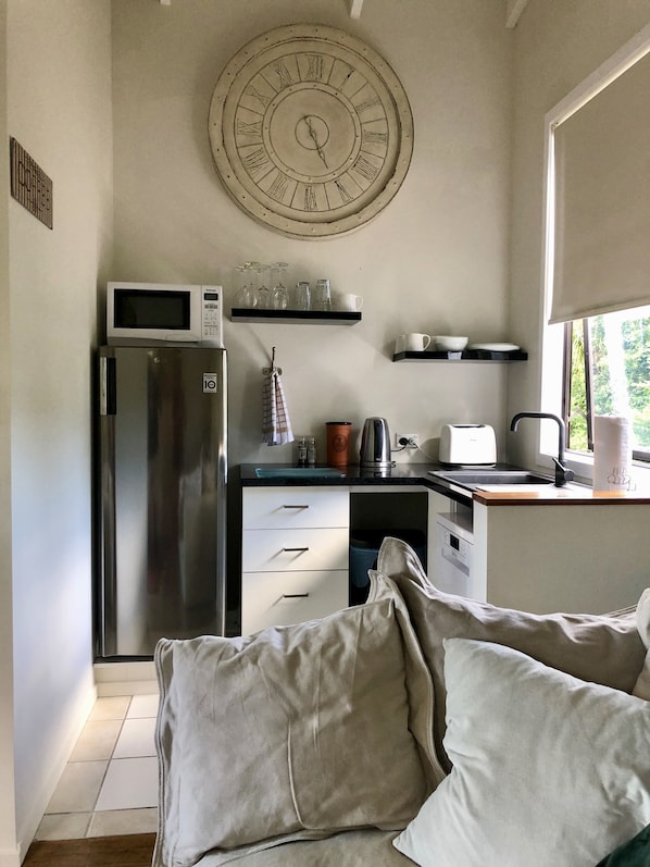 A modern kitchen with a stainless steel refrigerator, microwave, and kettle. There is a large wall clock above the counter, and a window providing natural light.