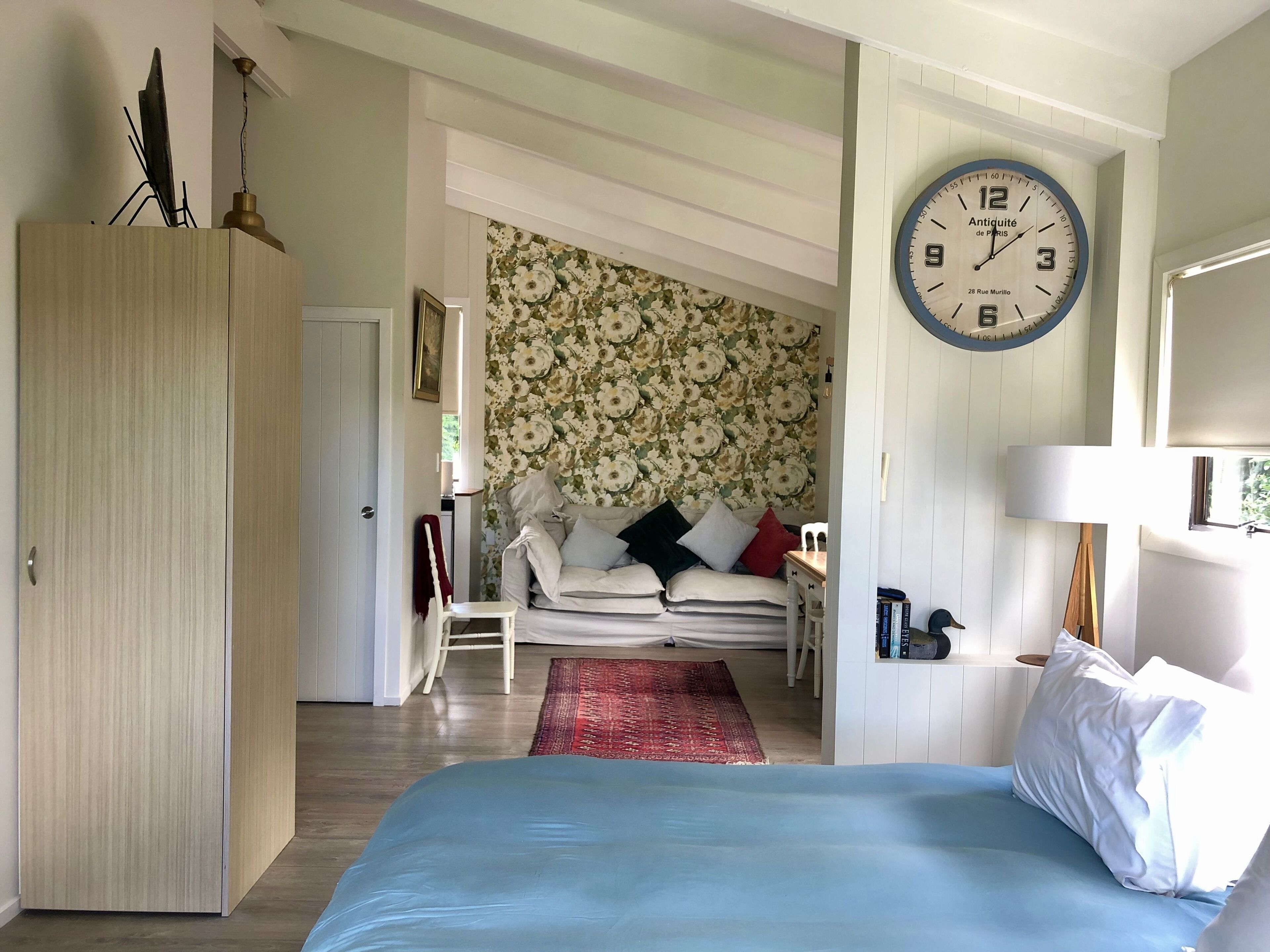 A bedroom with a bed, a wooden wardrobe, a floral wallpapered wall, a clock, and a red rug.