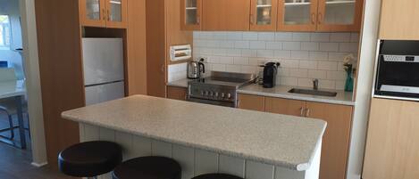 A modern kitchen with wooden cabinets, a central island with stools, a built-in oven, and a coffee maker.
