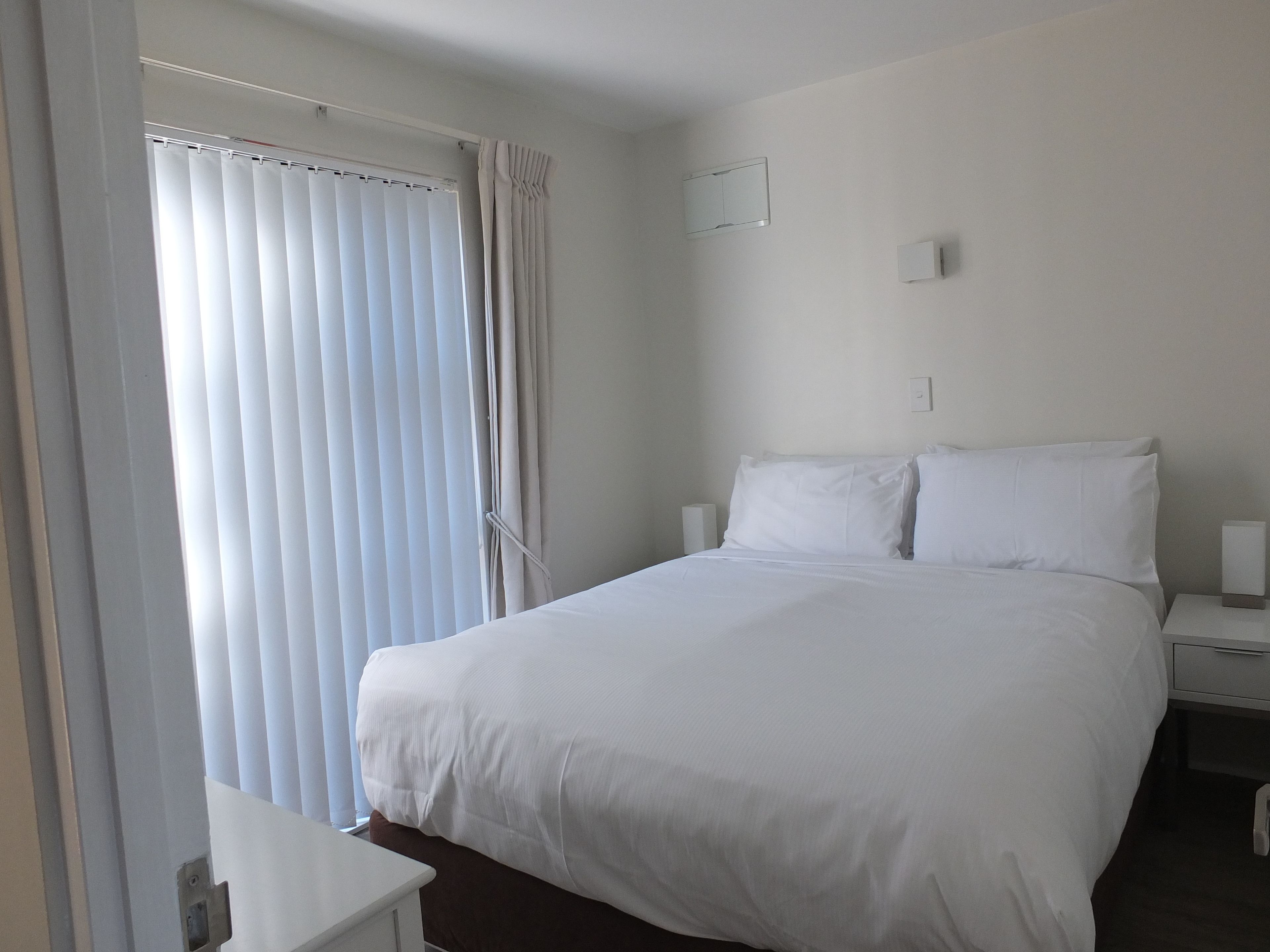 A hotel room with a large bed, a bedside table, a nightstand, and a window with blinds.