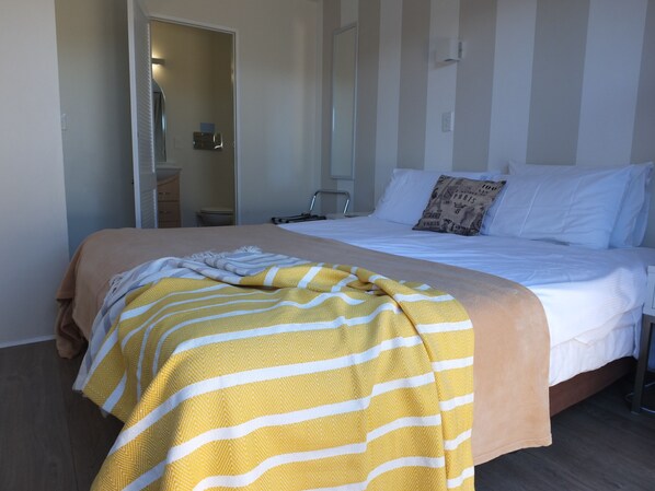 A hotel room with a bed, a bedside table, a lamp, and a door leading to a bathroom. - Le Chalet Waiheke Apartments (Waiheke Island)