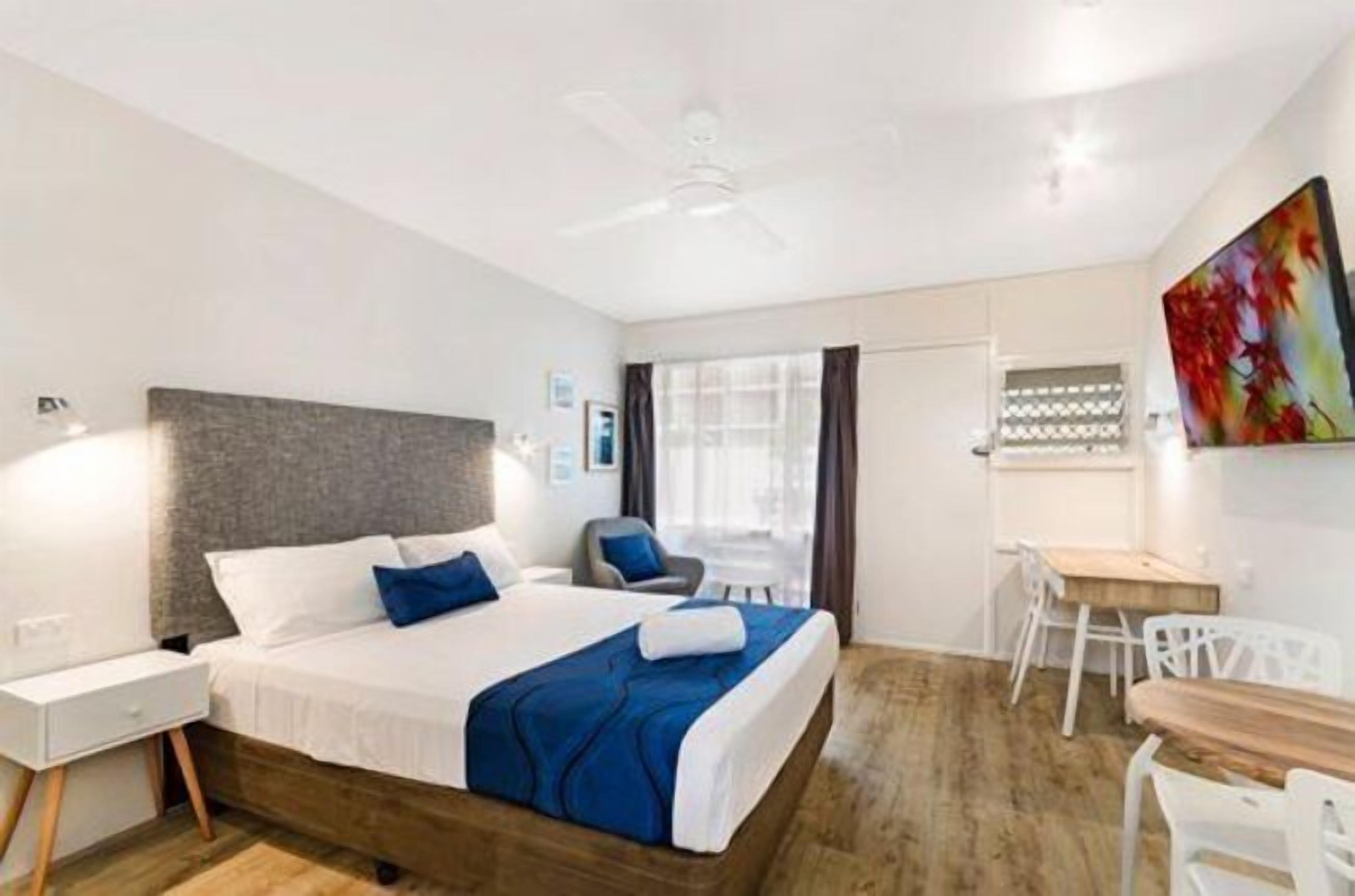 A bedroom with a bed, bedside table, chair, desk, and a painting on the wall.