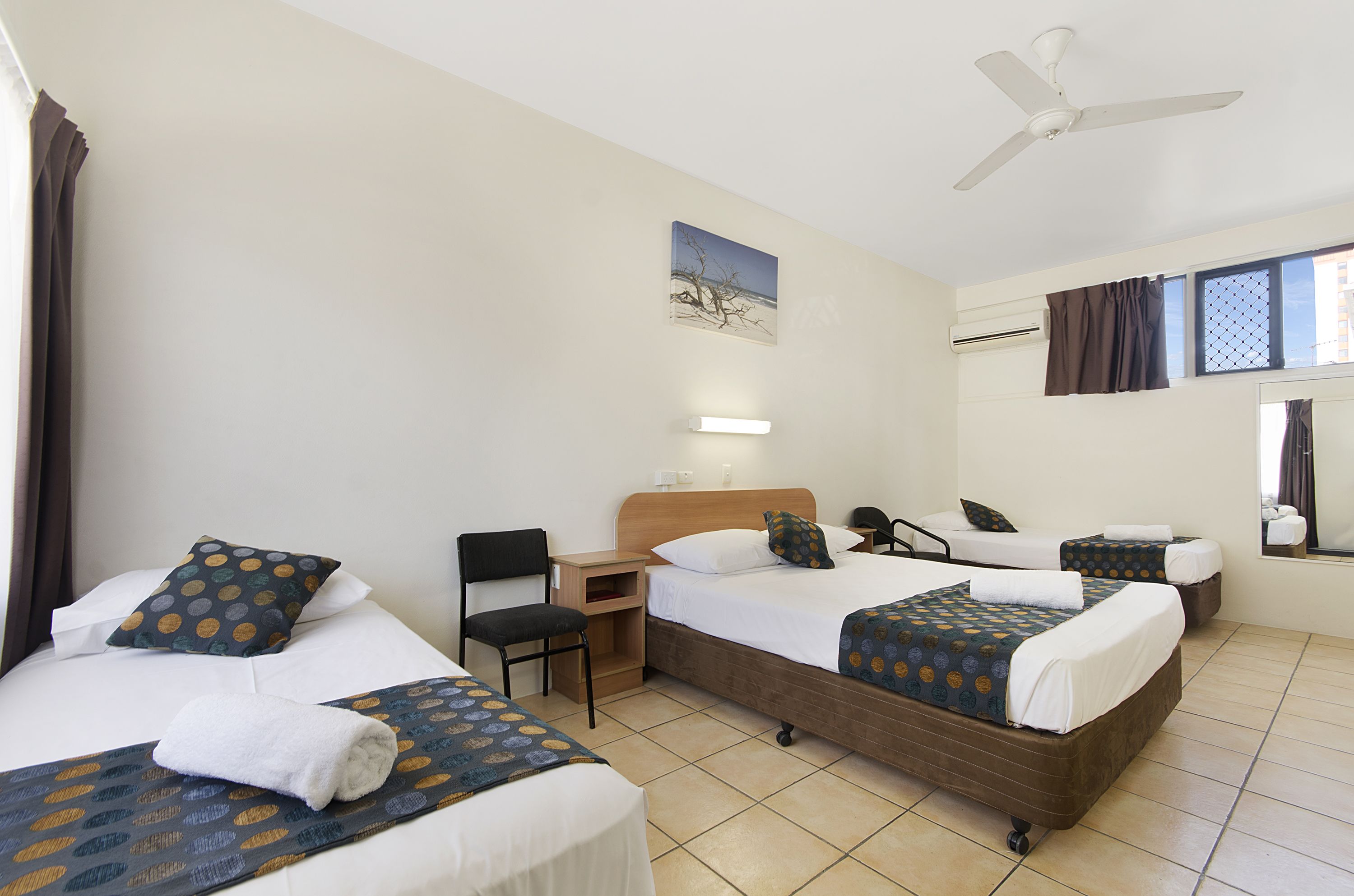 Ein Hotelzimmer mit drei Betten, einem Schreibtisch, einem Stuhl, einem Deckenventilator und einem Fenster mit Vorhängen.