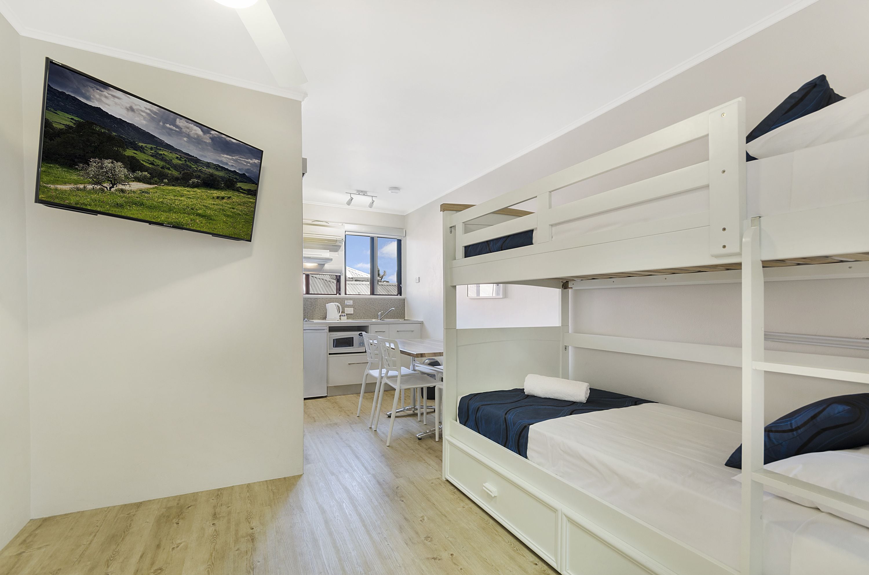 Une chambre avec des lits superposés, un bureau et une chaise, un tableau au mur et une vue sur la cuisine.
