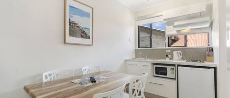 A compact kitchen with a wooden dining table, white chairs, a microwave, a refrigerator, and a window with a view of a building.