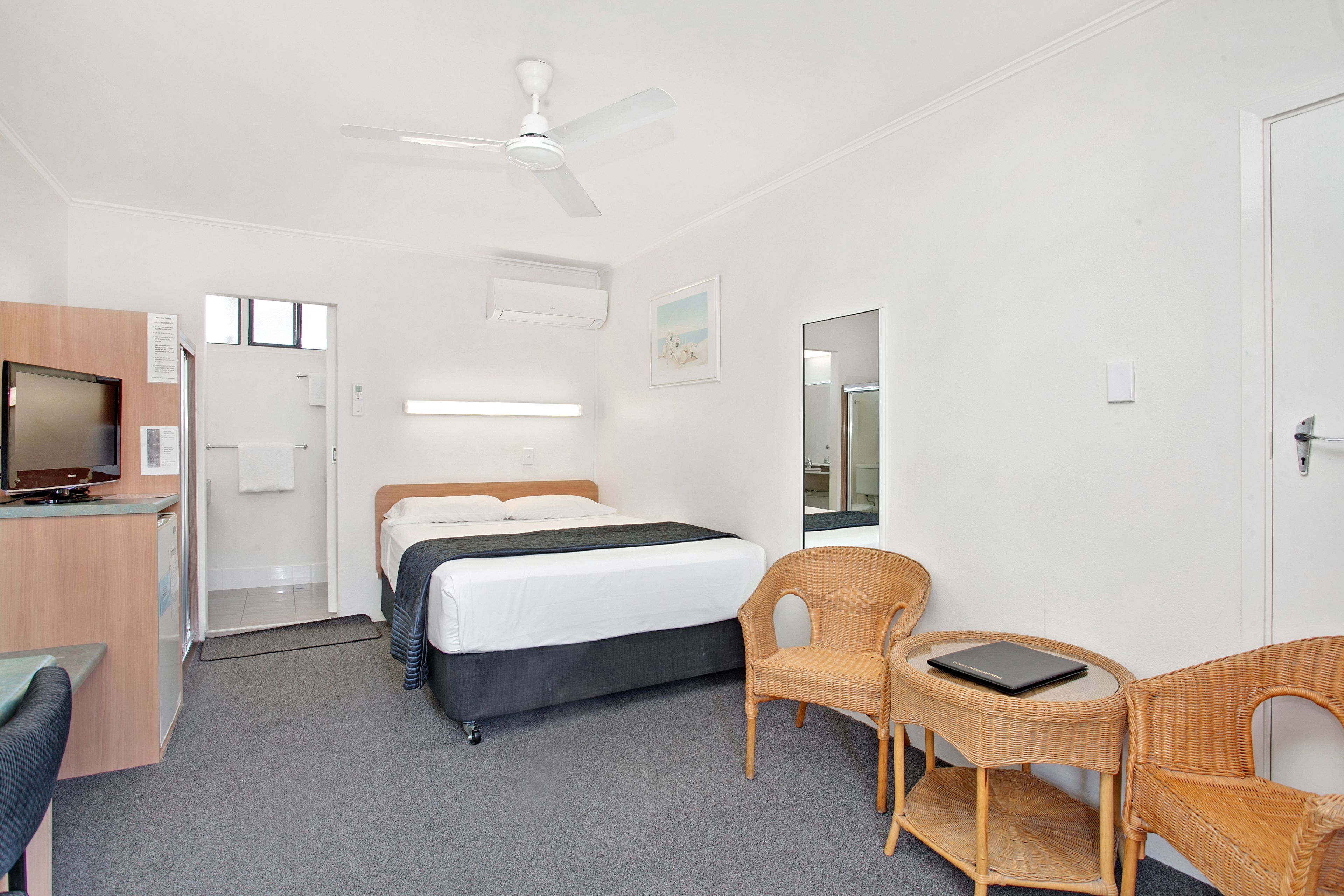 Una habitación de hotel con una cama, un escritorio con una computadora, dos sillas de mimbre, un ventilador de techo y un espejo grande.