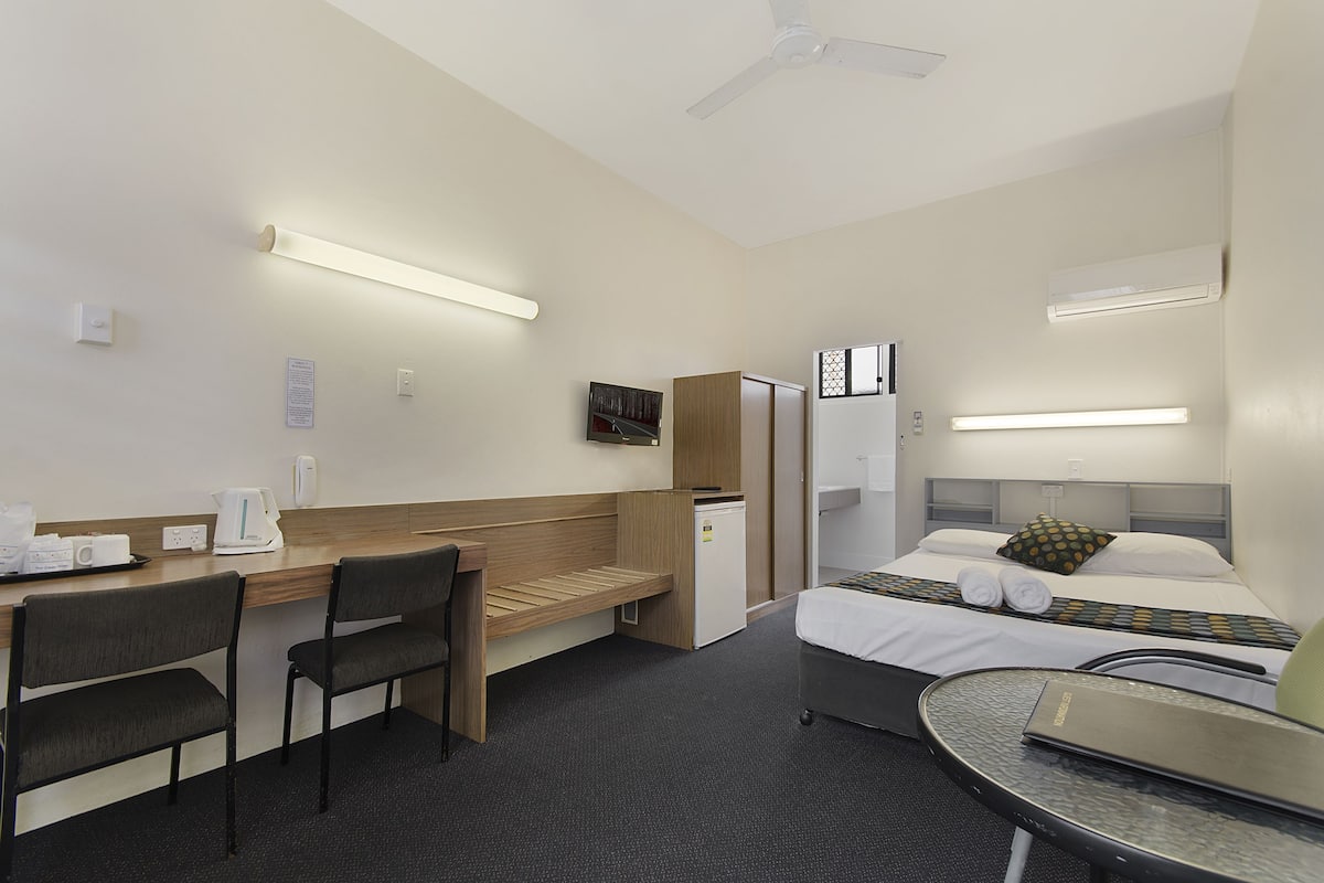 Une chambre d’hôtel équipée d’un lit, d’un bureau, de chaises, d’un petit réfrigérateur, d’une télévision et d’un ventilateur de plafond.