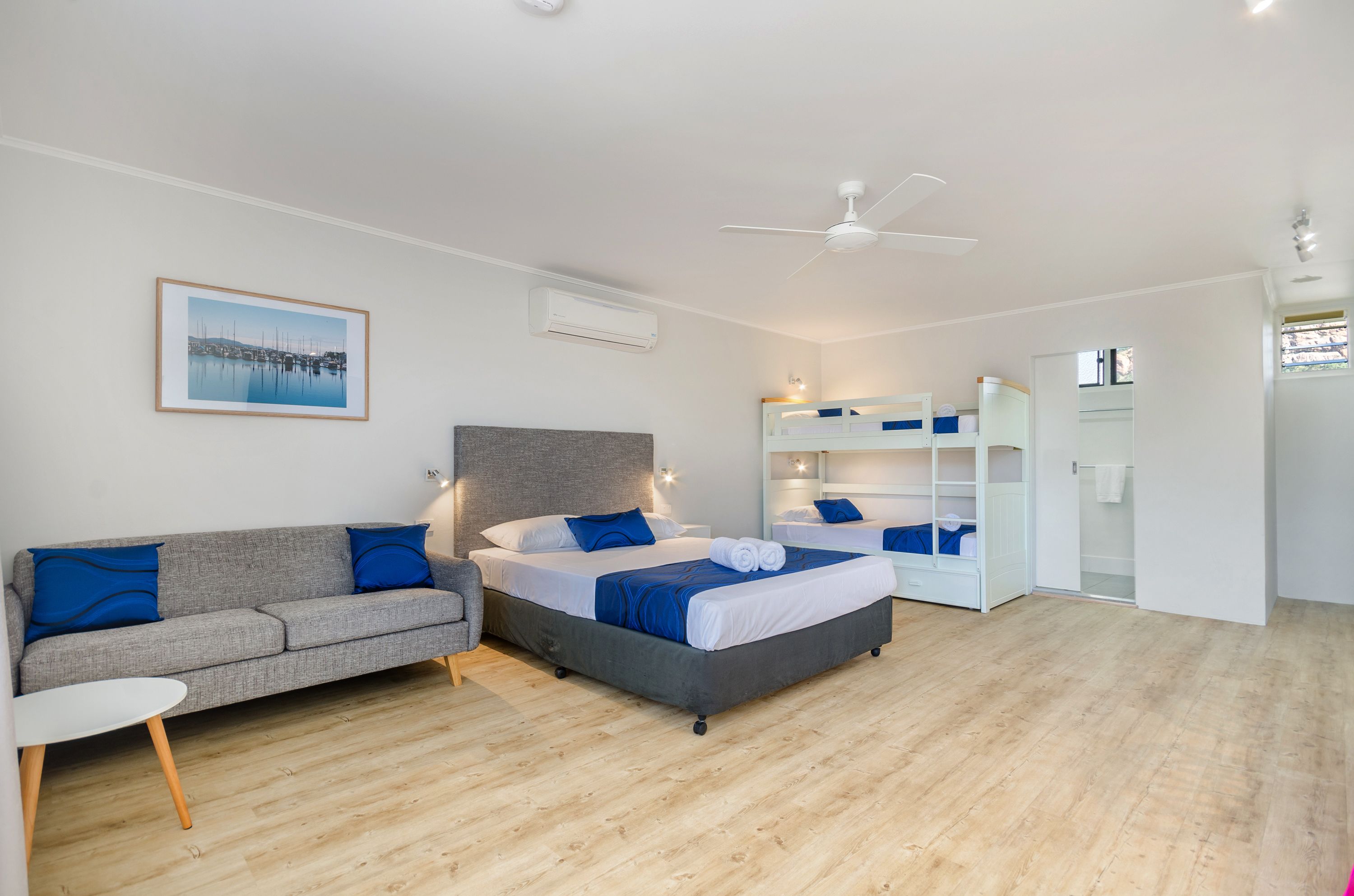 Ein modernes Schlafzimmer mit grauem Sofa, einem Bett mit blauer Bettwäsche, einem Deckenventilator und einem gerahmten Bild an der Wand.