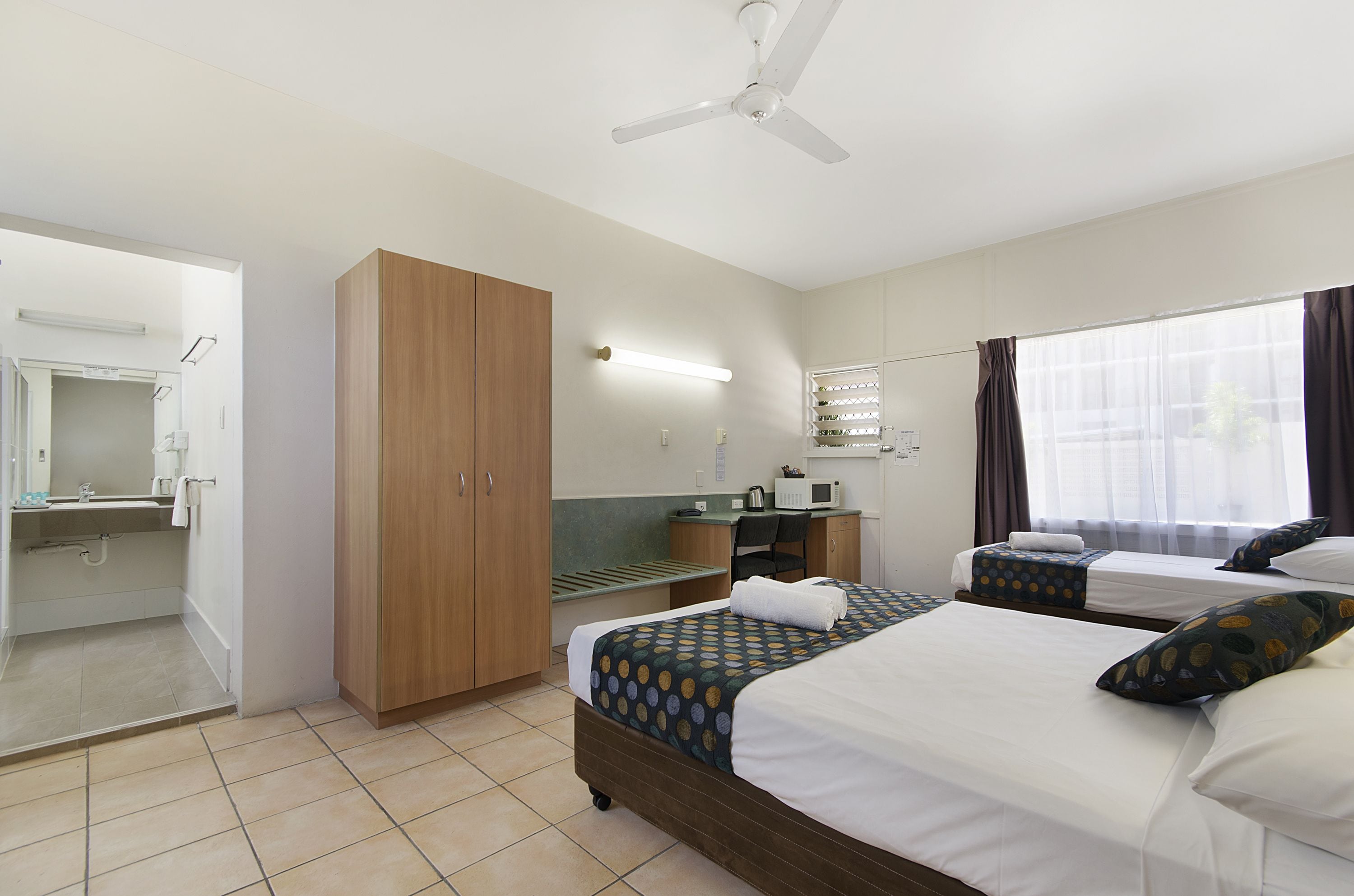 A hotel room with two beds, a desk, a mirror, and a ceiling fan.