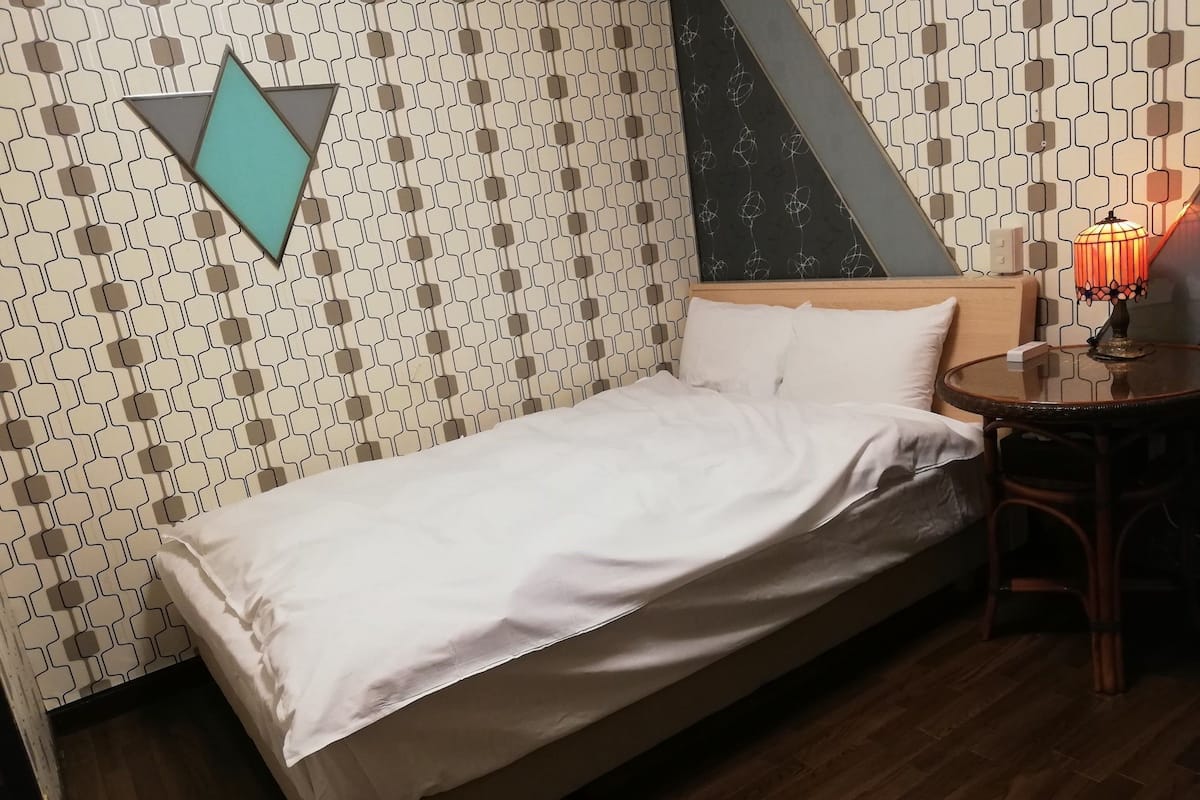A single bed with a white sheet, a wooden headboard, a patterned wall, a small round table with a lamp, and a wooden floor.