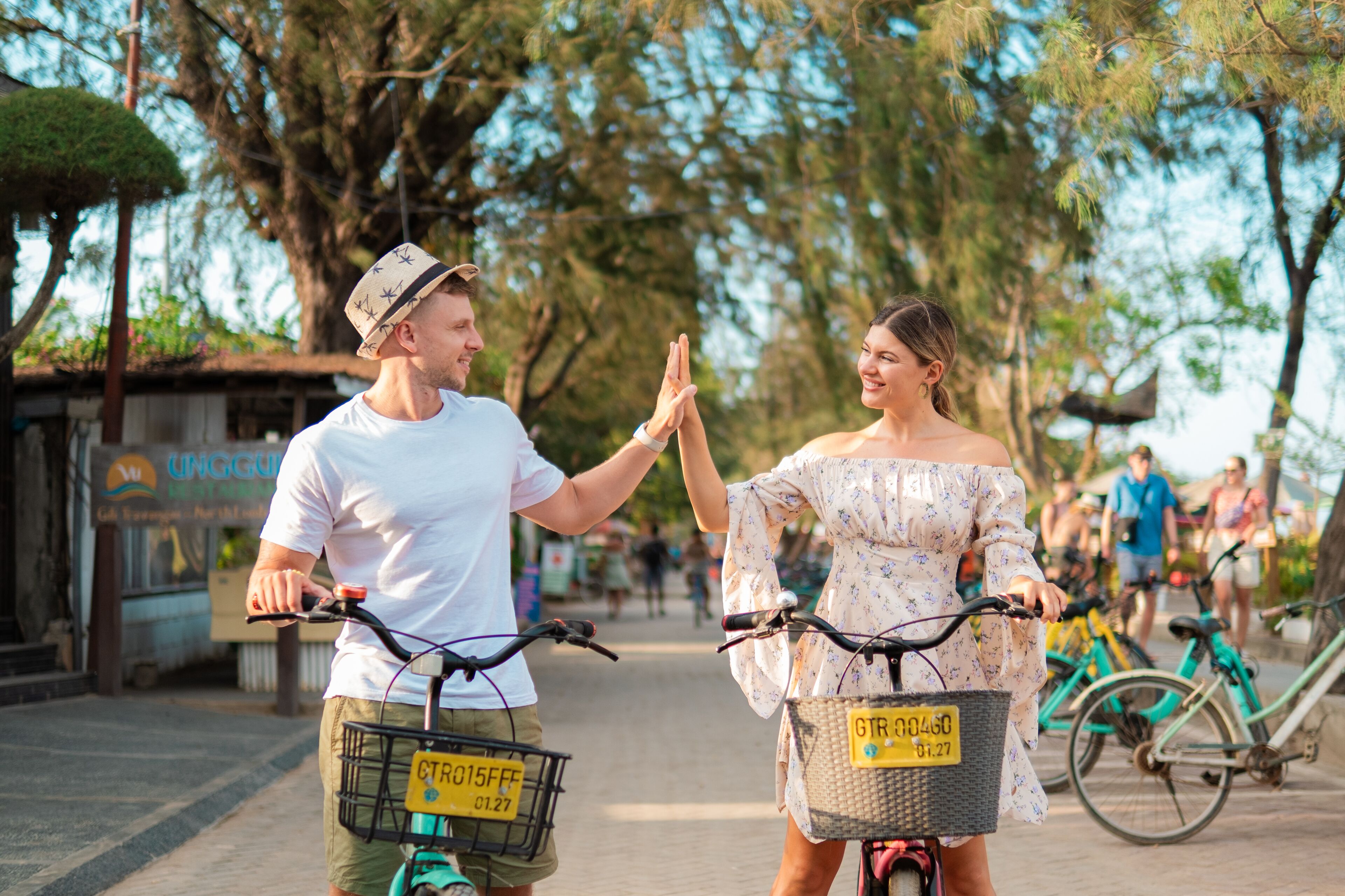 Twee personen op fietsen, één met een wit t-shirt en hoed, de ander in een bloemenjurk, met een straat en geparkeerde fietsen op de achtergrond.