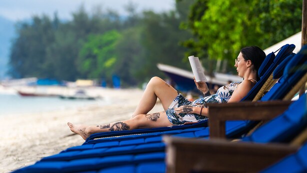A person relaxing on a blue lounge chair with a beach and boats in the background. - Villa Almarik (Gili Trawangan)