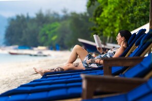 A person relaxing on a blue lounge chair with a beach and boats in the background. - Lotus Almarik Gili (Gili Trawangan)
