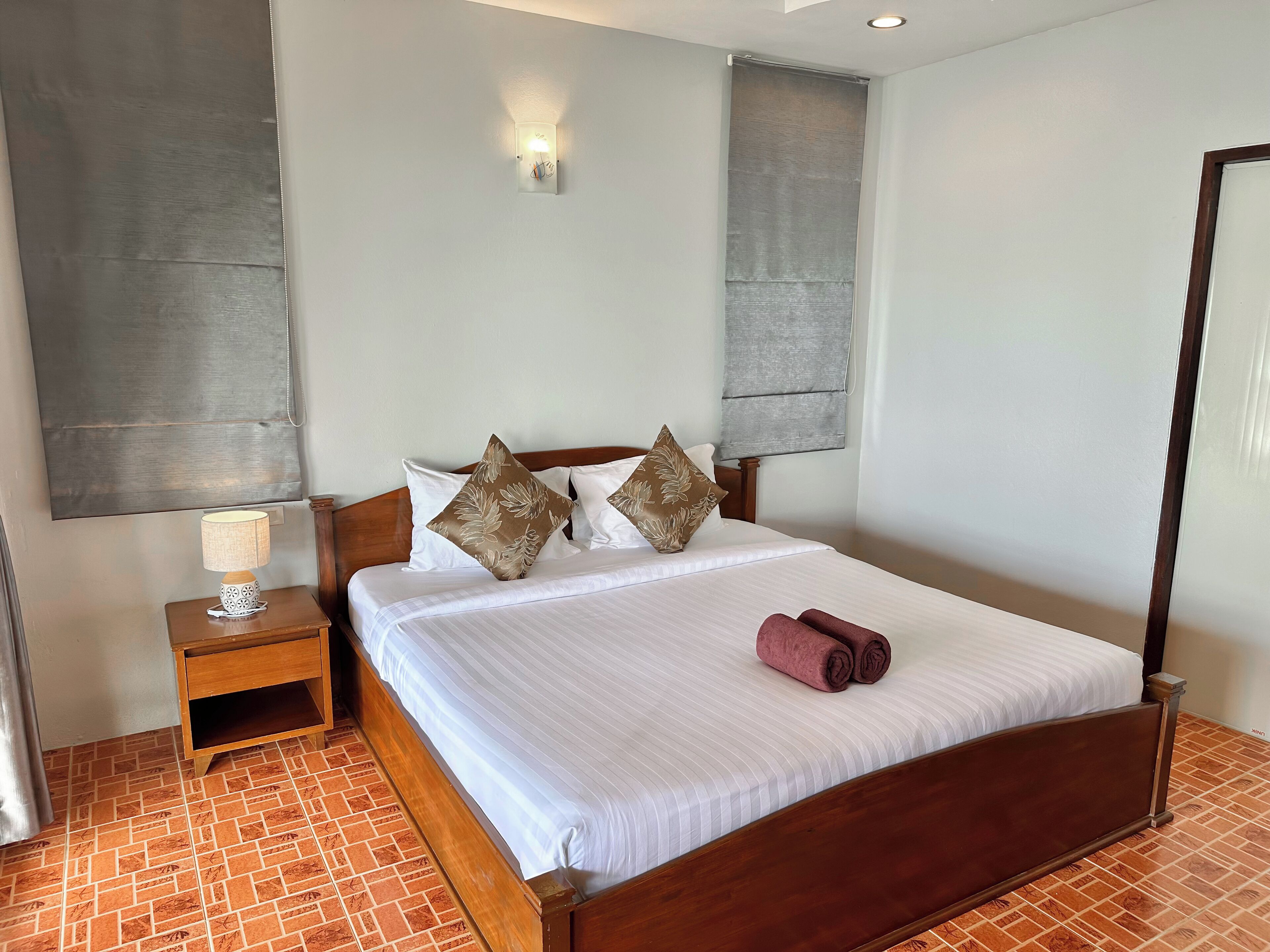 A neatly made bed with white linens and decorative pillows, a wooden nightstand with a lamp, and a brick-patterned floor.