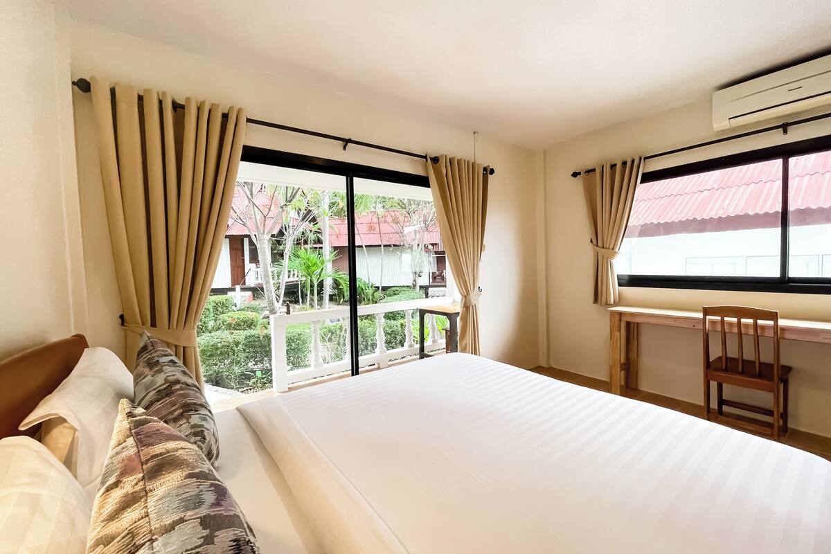 Habitación de hotel con una cama grande, una silla de madera y un escritorio. Hay una ventana con cortinas, y se ve un edificio y follaje afuera.