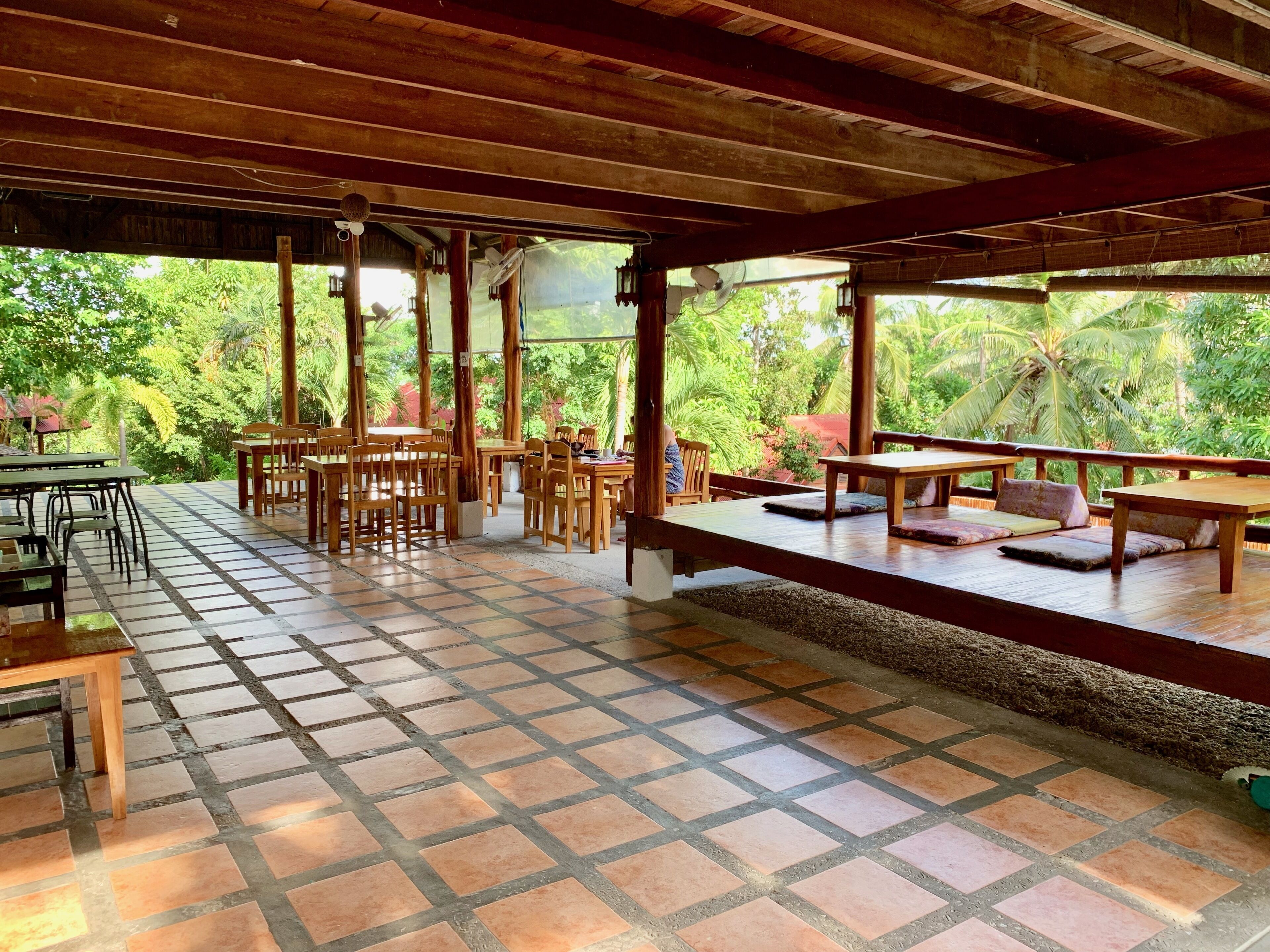 Uma área de refeições ao ar livre com mesas e cadeiras de madeira, cercada por uma pérgola de madeira e vista para vegetação exuberante.