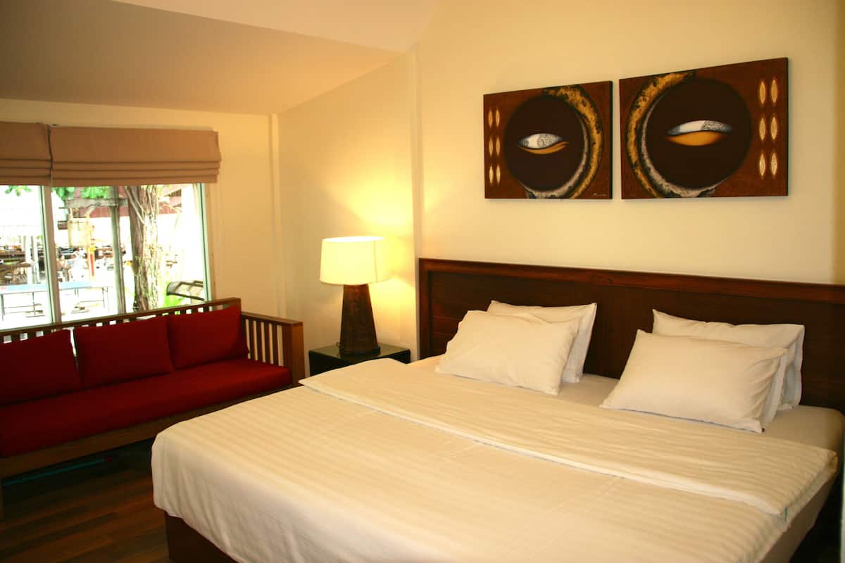 A hotel room with a bed, a red sofa, a nightstand with a lamp, and two wall art pieces.