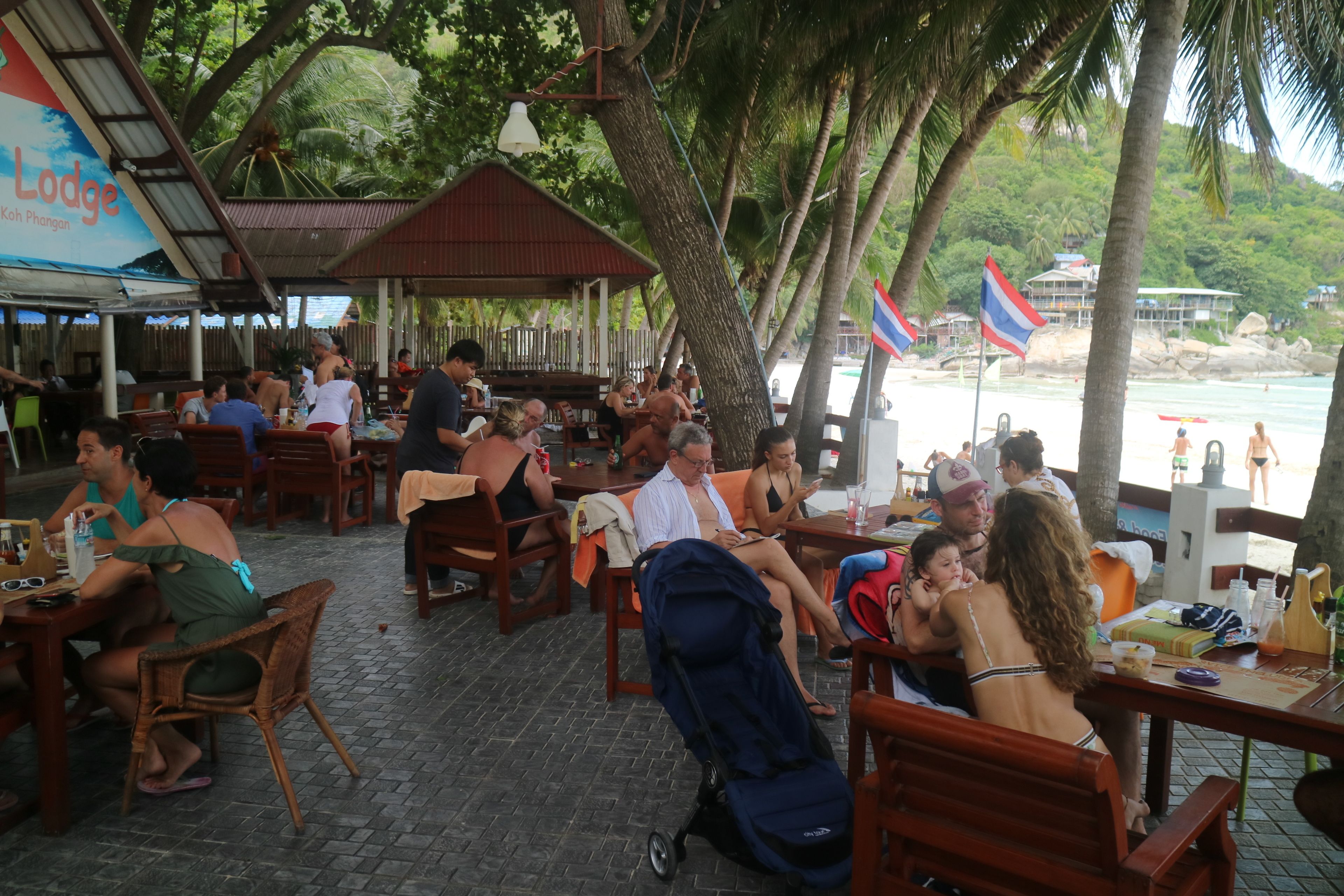 An outdoor dining area with wooden tables and chairs, surrounded by palm trees and a view of the beach.
