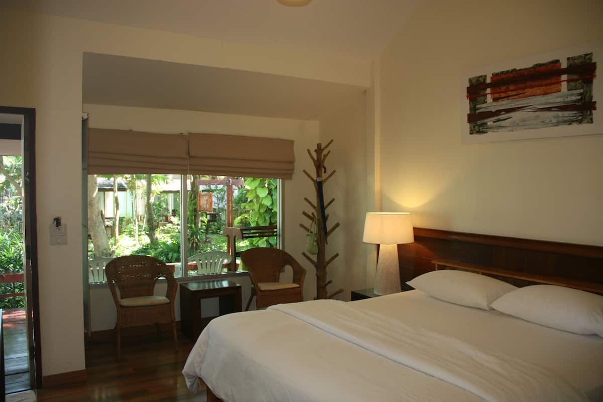 A hotel room with a bed, a chair, a table, and a window with a view of greenery.